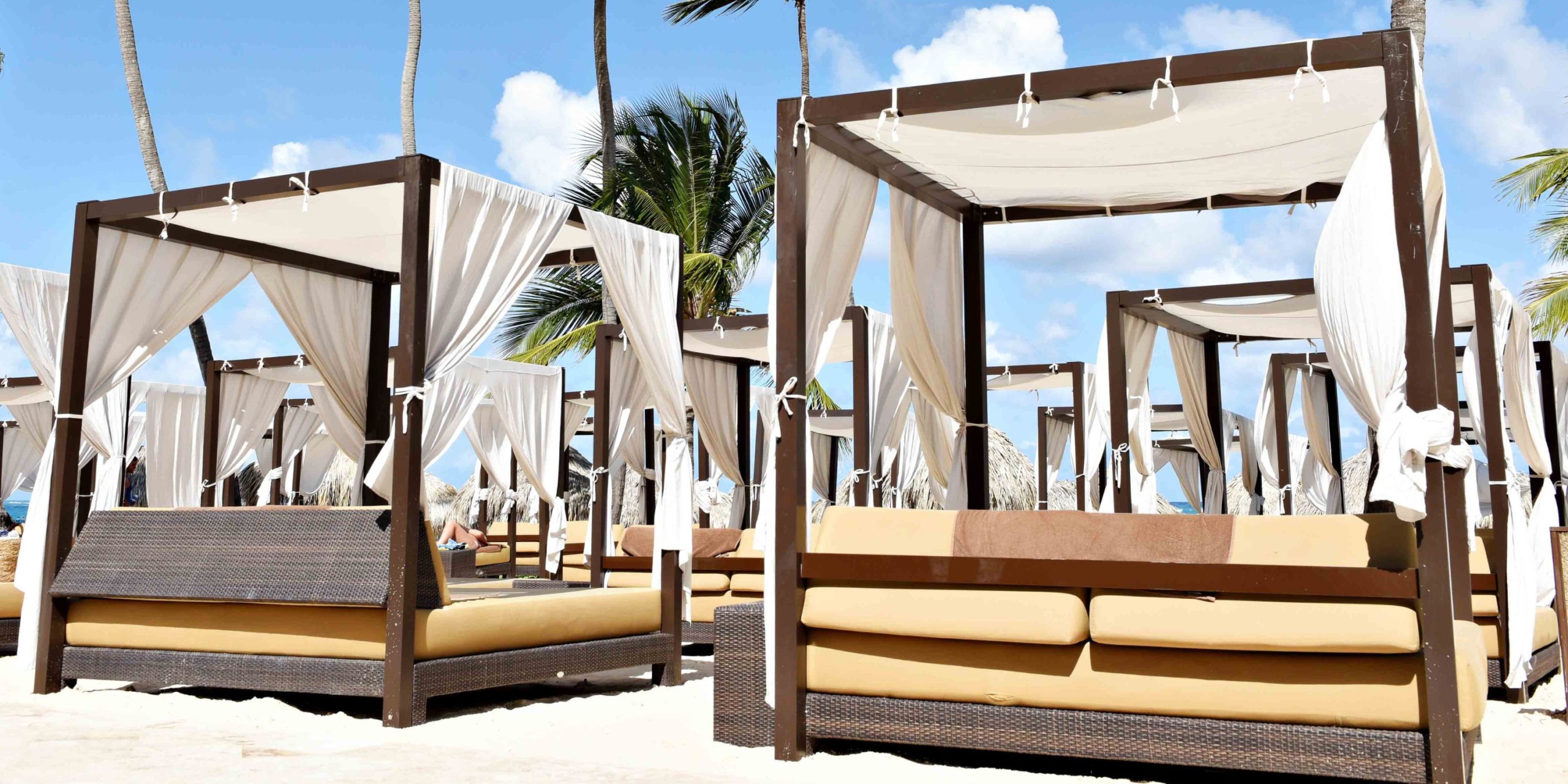 The Royalton Punta Cana features outdoor cabanas adorned with white curtains and beige cushions, set against the backdrop of a sunny beach with majestic palm trees.