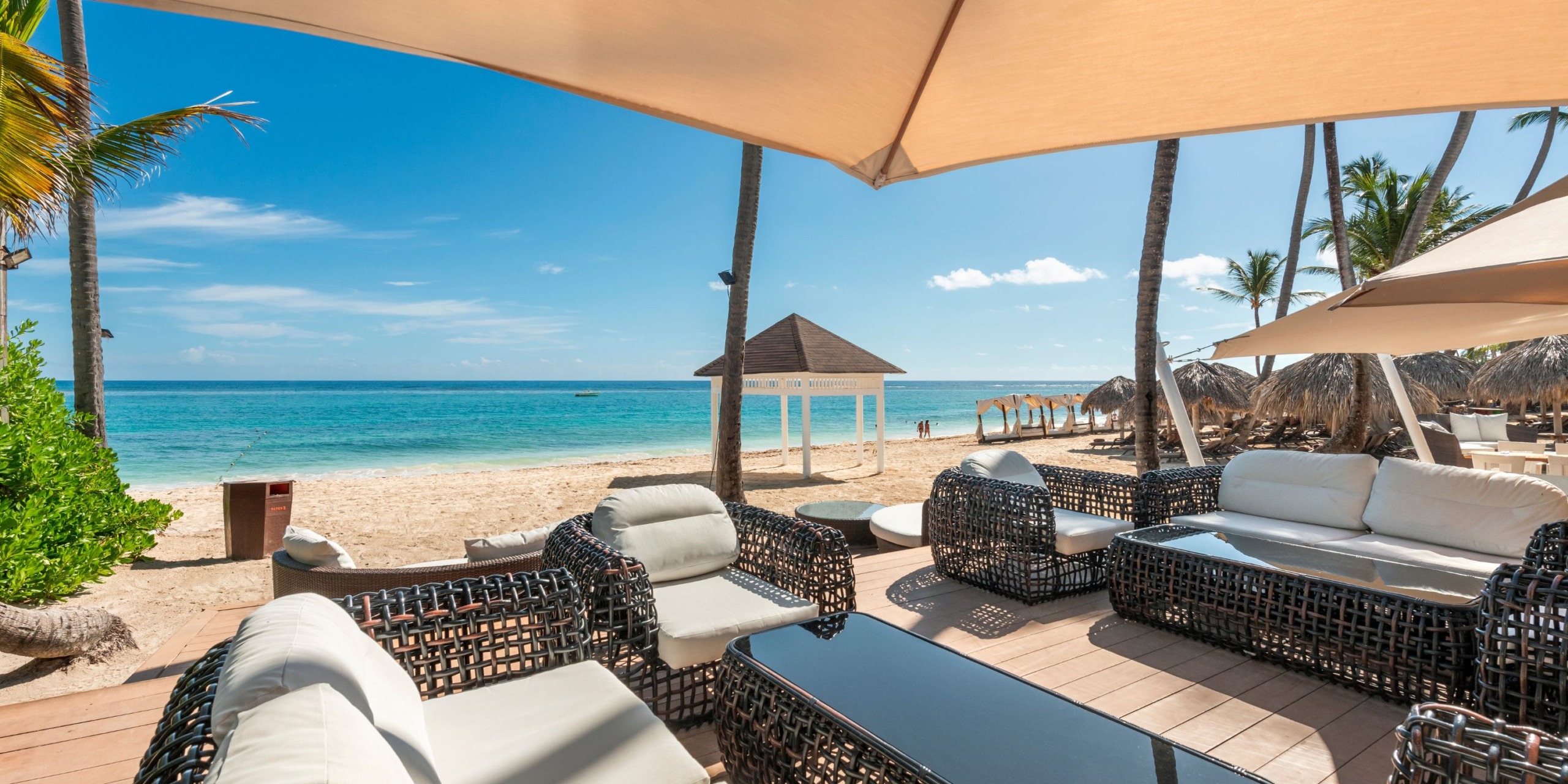The patio at Royalton Punta Cana offers wicker furniture with a stunning view of a sandy beach and turquoise ocean, shaded by large umbrellas, with palm trees and huts visible in the background.