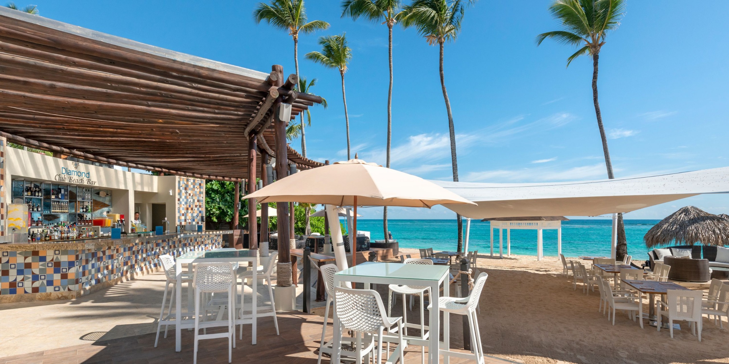At the Royalton Punta Cana, enjoy an outdoor beach bar with high tables and chairs under umbrellas, surrounded by palm trees, while the ocean shimmers in the background under a clear blue sky.
