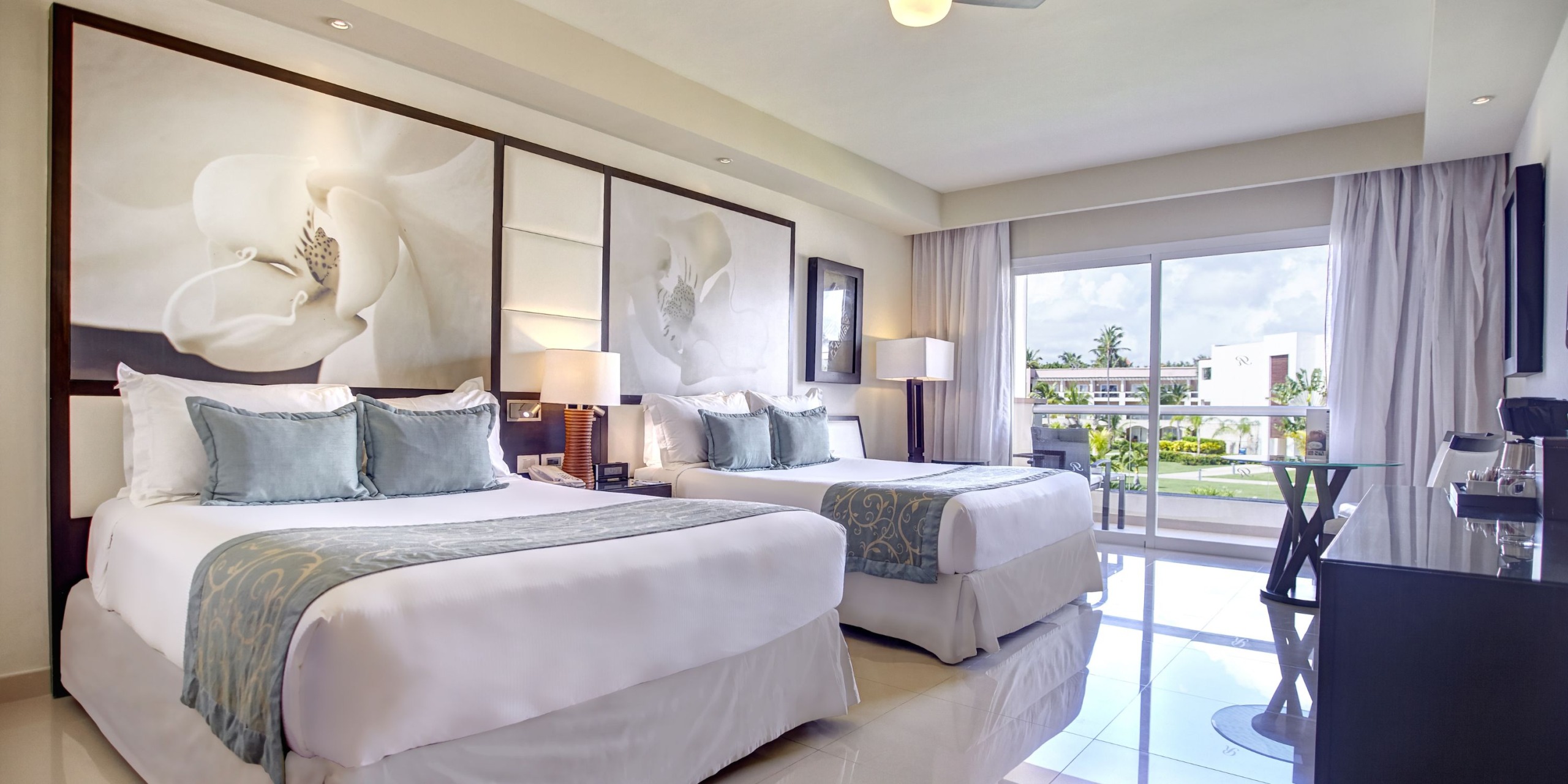 Experience a stay at the Royalton Punta Cana with a hotel room featuring two double beds, decorative pillows, large floral wall art, a desk, and a balcony offering views of lush greenery and city buildings.