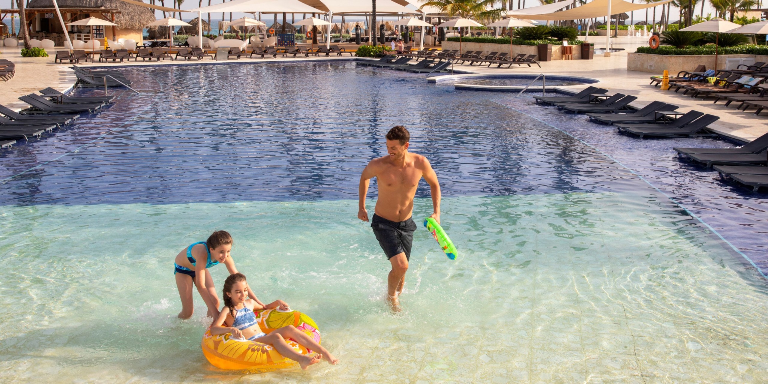 A man and two children are enjoying the pool at Royalton Punta Cana. One child is relaxing on an inflatable ring while the others wade through the shallow water. Lounge chairs can be seen in the background.