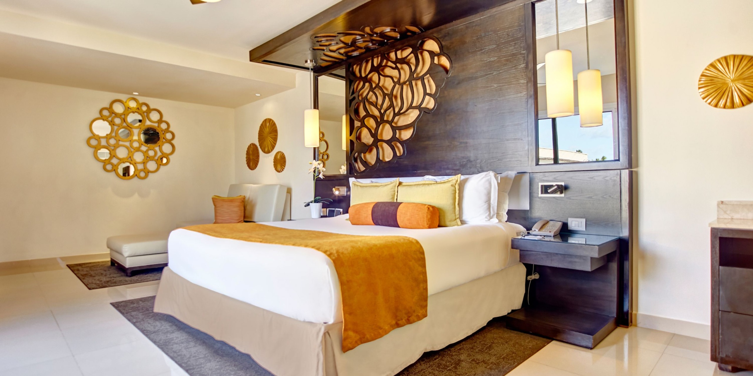 Experience the Royalton Punta Cana, featuring a modern hotel room with a king-sized bed, decorative wall art, pendant lights, and a cushioned bench. Warm tones and elegant decor come together to create a cozy atmosphere.