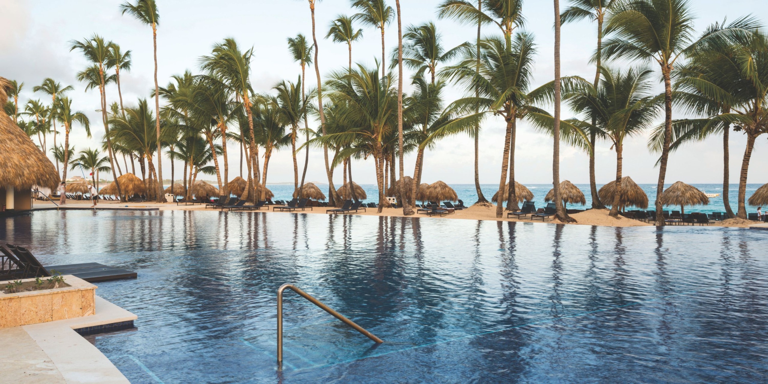 The infinity pool at Royalton Punta Cana features a metal handrail and offers breathtaking views of a sandy beach adorned with palm trees and thatched umbrellas.