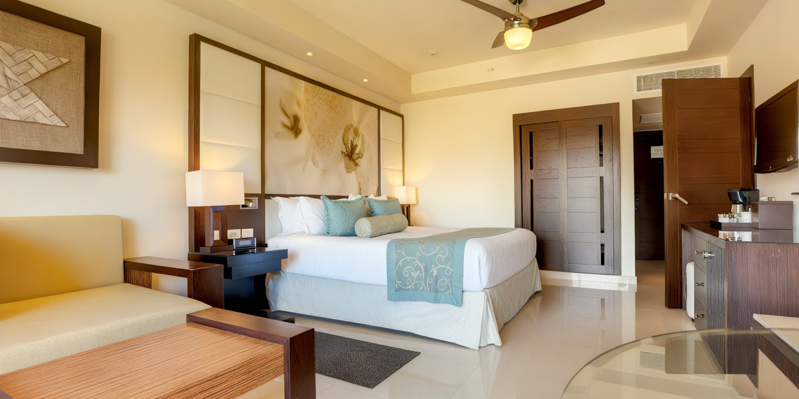 Room in the Royalton Punta Cana featuring a neatly made bed adorned with decorative pillows and modern furnishings. The setting is completed by a ceiling fan, wall art, and lamps. Light floods in from an unseen window.