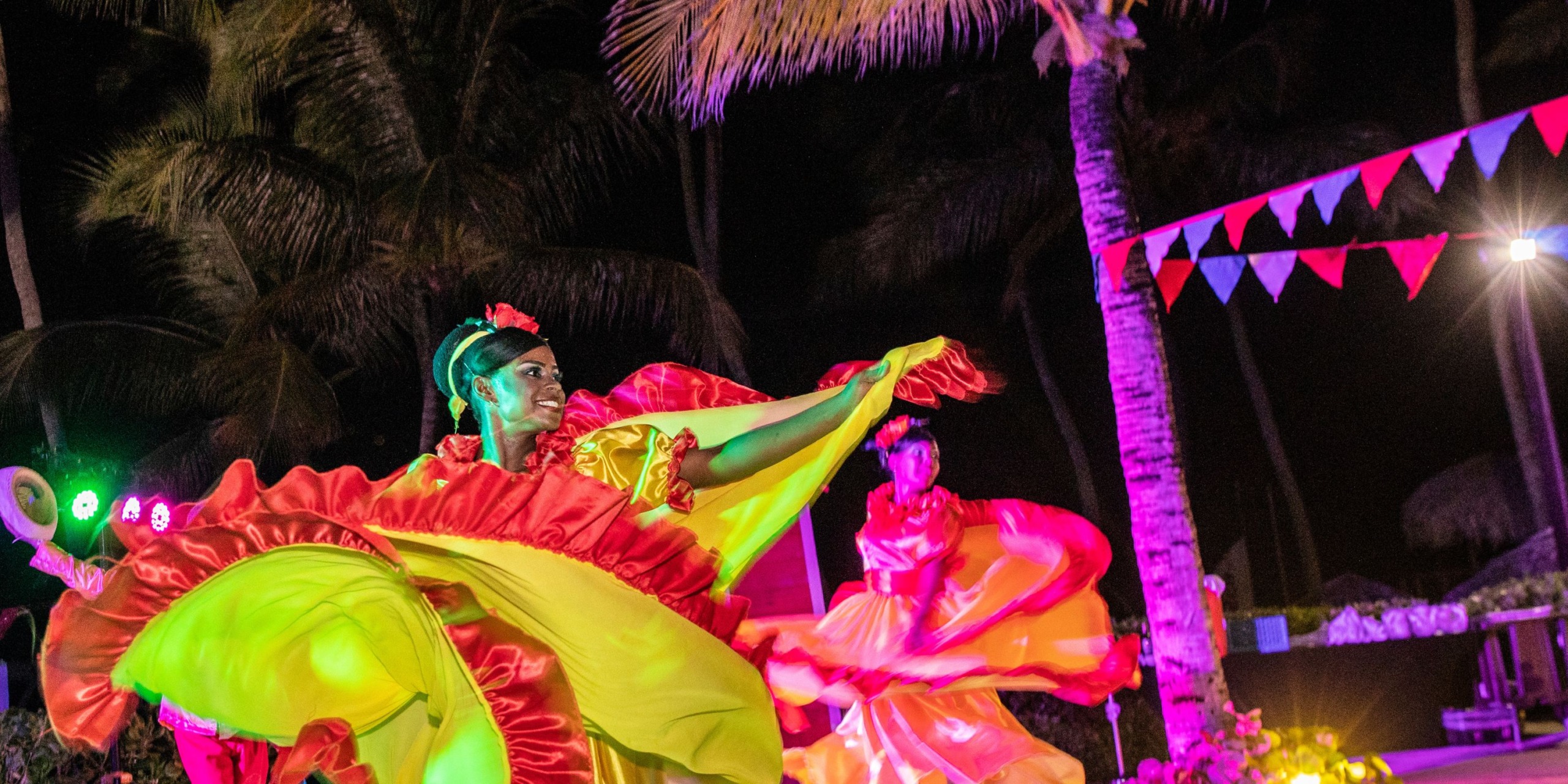 Under the enchanting night sky of Royalton Punta Cana, two dancers in vibrant costumes perform amidst the palm trees, surrounded by colorful lights and festive bunting.