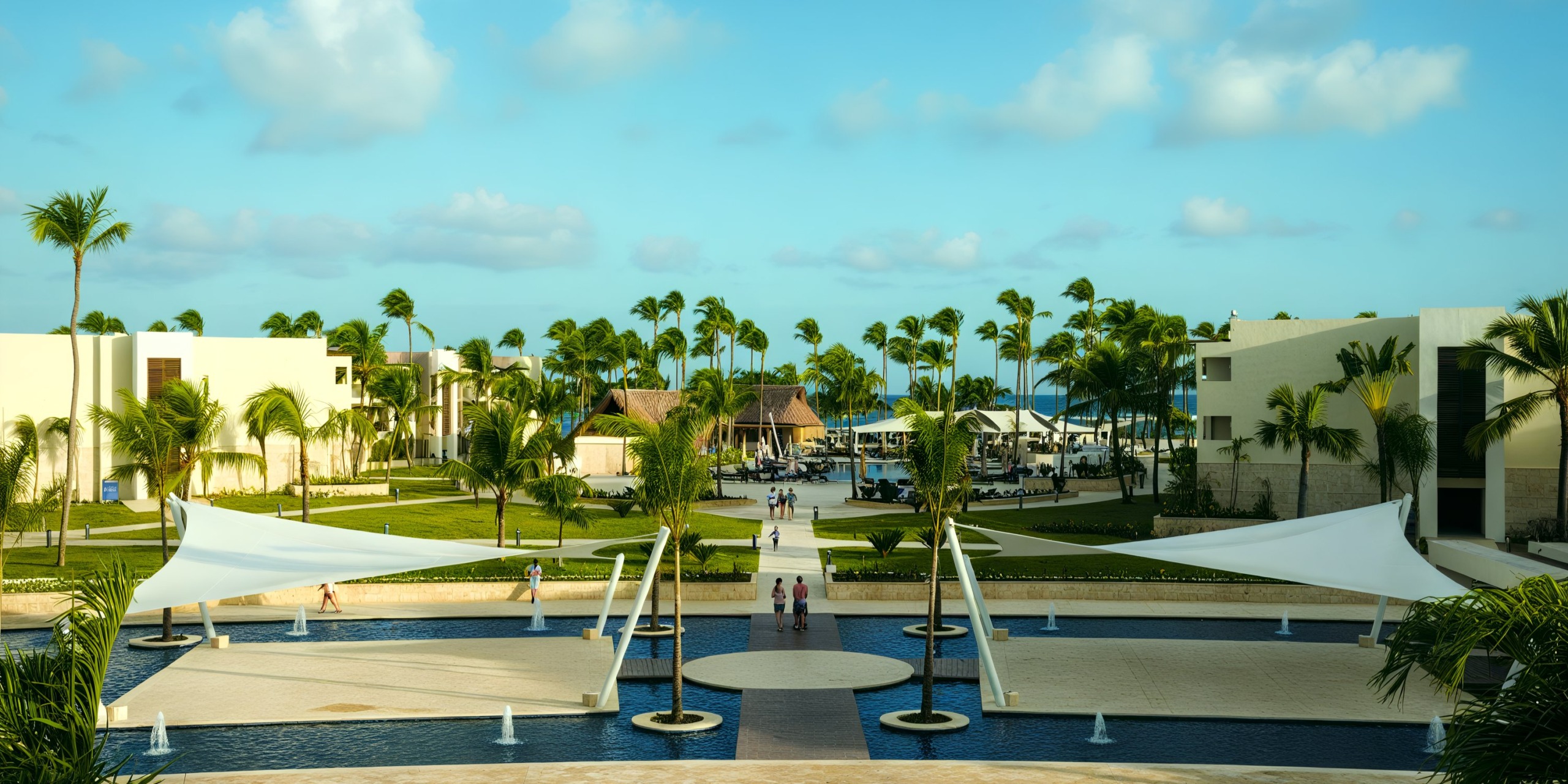 The Royalton Punta Cana features a resort view with palm trees, modern buildings, and a central pool area shaded by white sails under a blue sky.