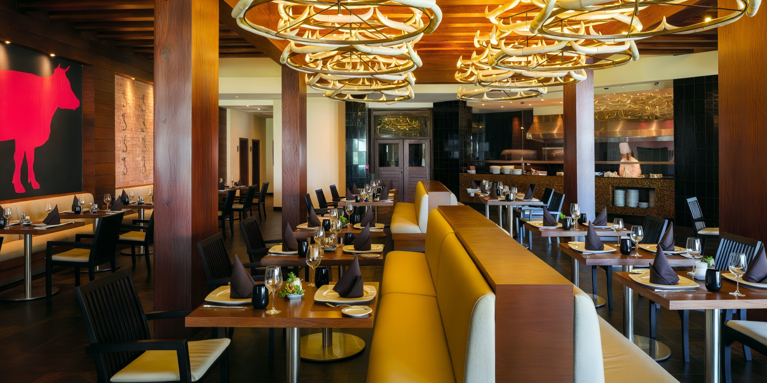 The Royalton Punta Cana boasts a contemporary restaurant interior with wooden beams and vibrant yellow seating. The space features an impressive large red cow artwork adorning the wall, complemented by elegant chandeliers gracefully suspended from the ceiling. Tables are meticulously set for dining.