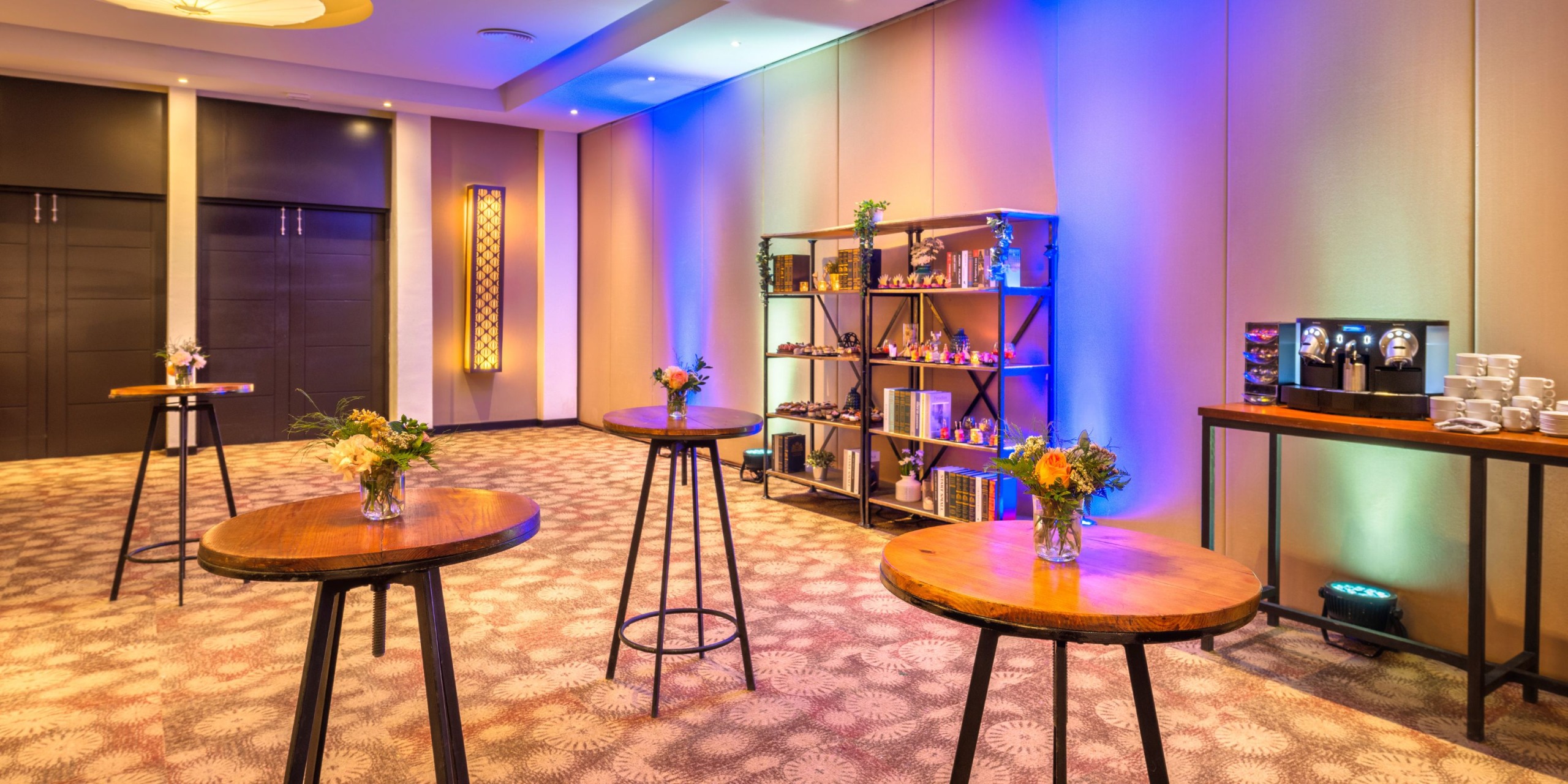 The Royalton Punta Cana features a brightly lit room adorned with small round tables, each decorated with flowers. Shelves displaying various items and a digital screen are set against a vibrant and colorful wall.