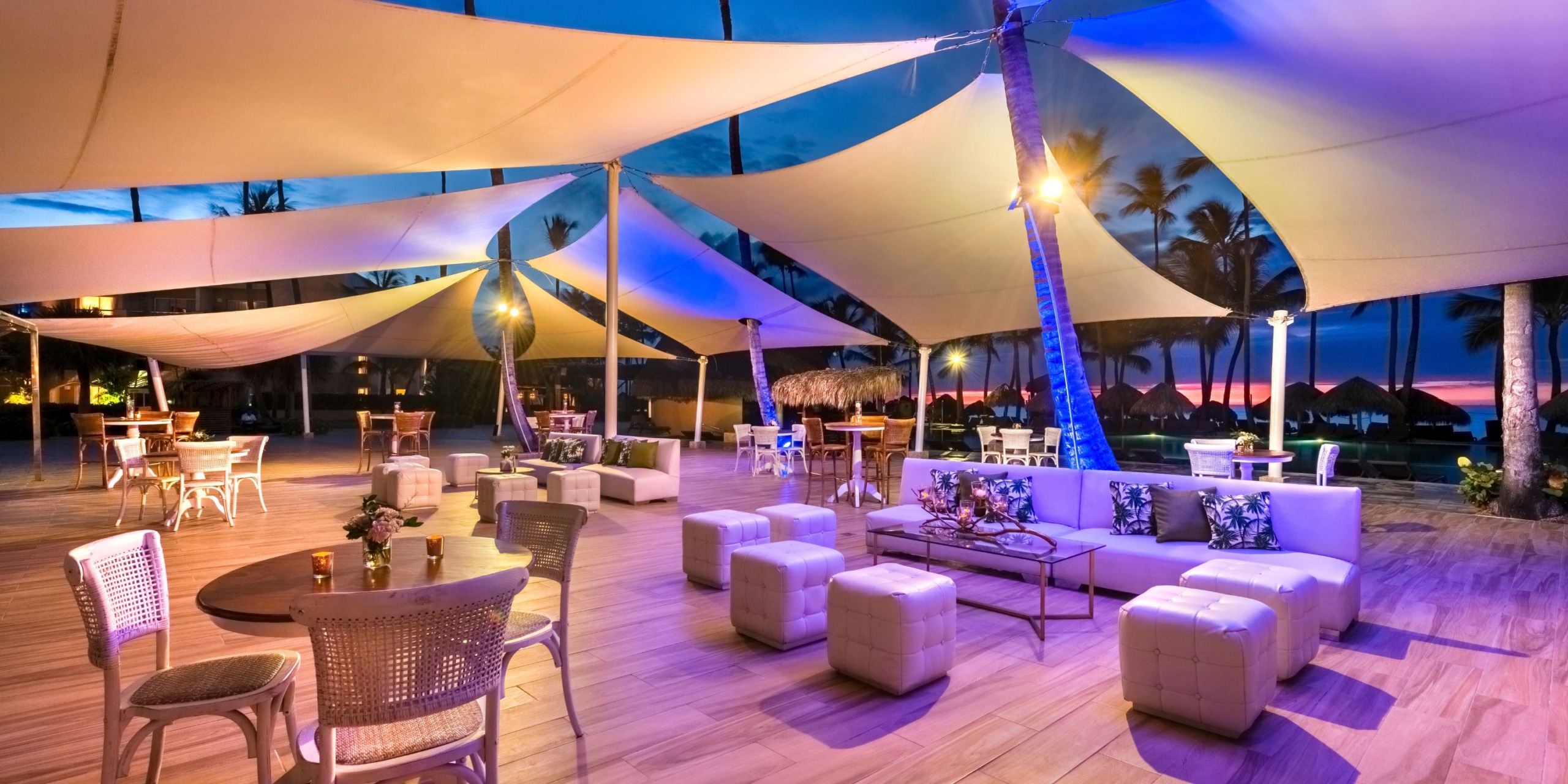 The Royalton Punta Cana features an outdoor patio adorned with elegant white furniture and large fabric canopies, illuminated by vibrant, colorful lights. A variety of tables and chairs are stylishly arranged across the wooden floor, set against the backdrop of a breathtaking twilight sky.