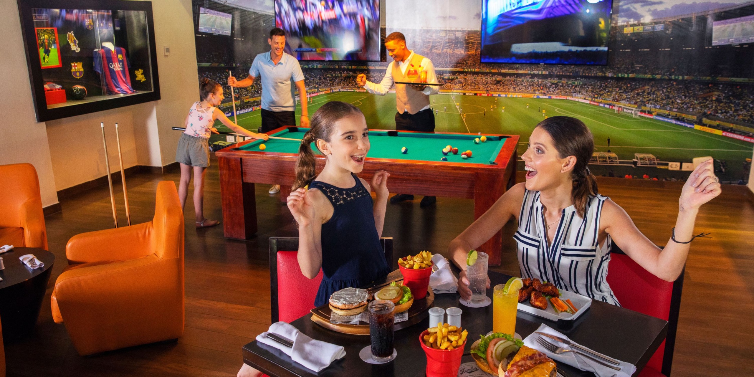 Guests enjoying their time at the Royalton Punta Cana, surrounded by a sports bar atmosphere with a pool table, expansive screens displaying a soccer match, and delicious meals laid out before them.