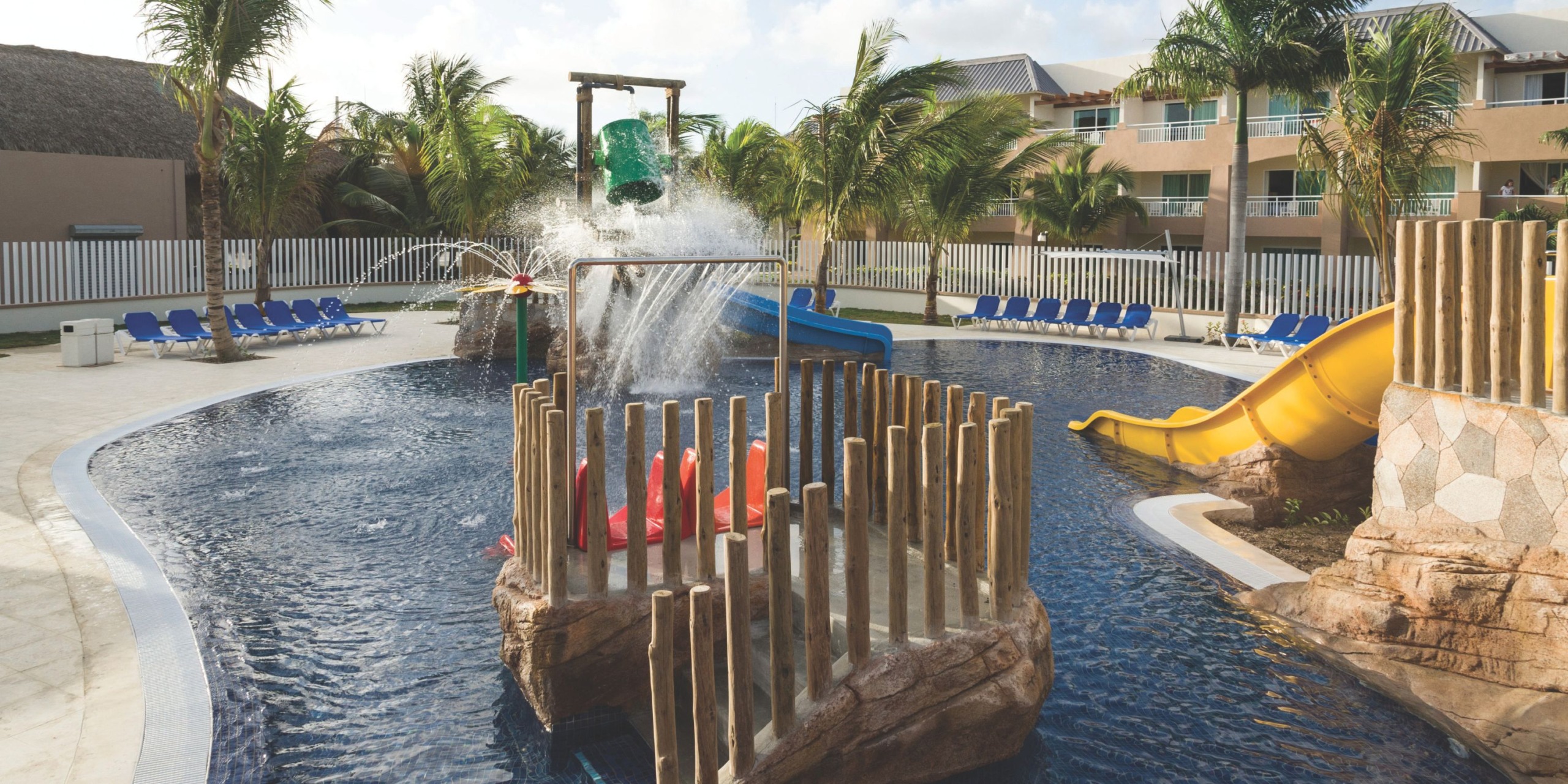 The Royalton Punta Cana features a resort pool water play area complete with slides, a water bucket, and lounge chairs, all surrounded by palm trees and a fence.