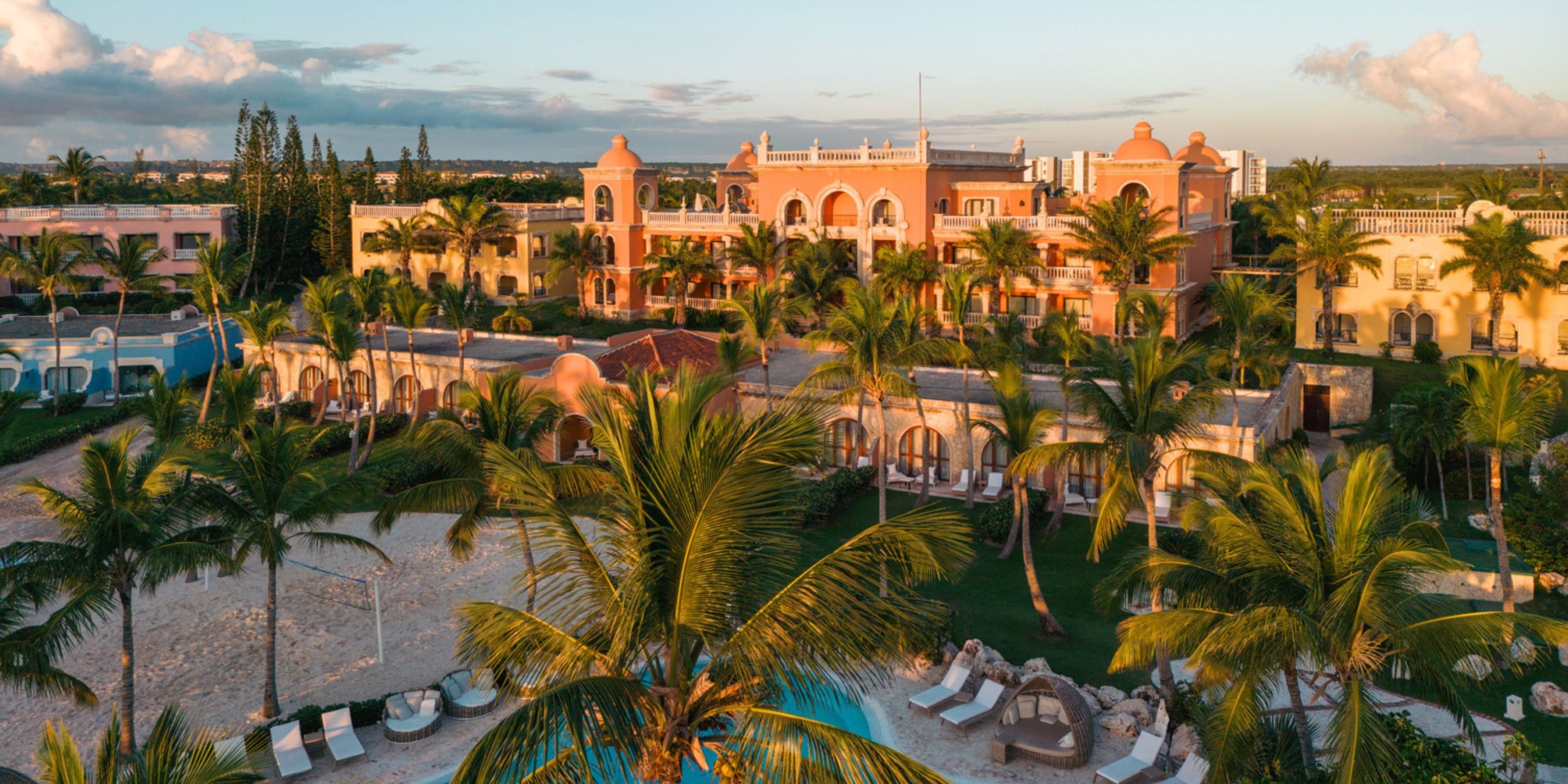 Aerial view of Sanctuary Cap Cana, a tropical beachfront resort featuring a large pink building, surrounded by palm trees, pools, and lounge areas under a clear sky.