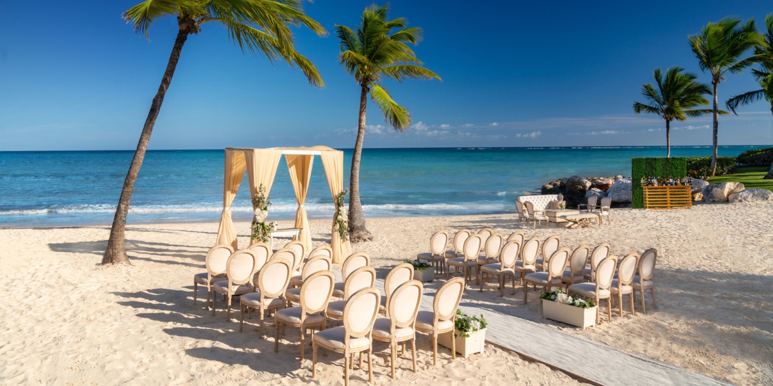 Sanctuary Cap Cana offers a stunning beach wedding setup, featuring rows of chairs directed towards an altar beneath a beautiful canopy. The scene is elegantly framed by palm trees, with the majestic ocean serving as a backdrop under the clear blue sky.