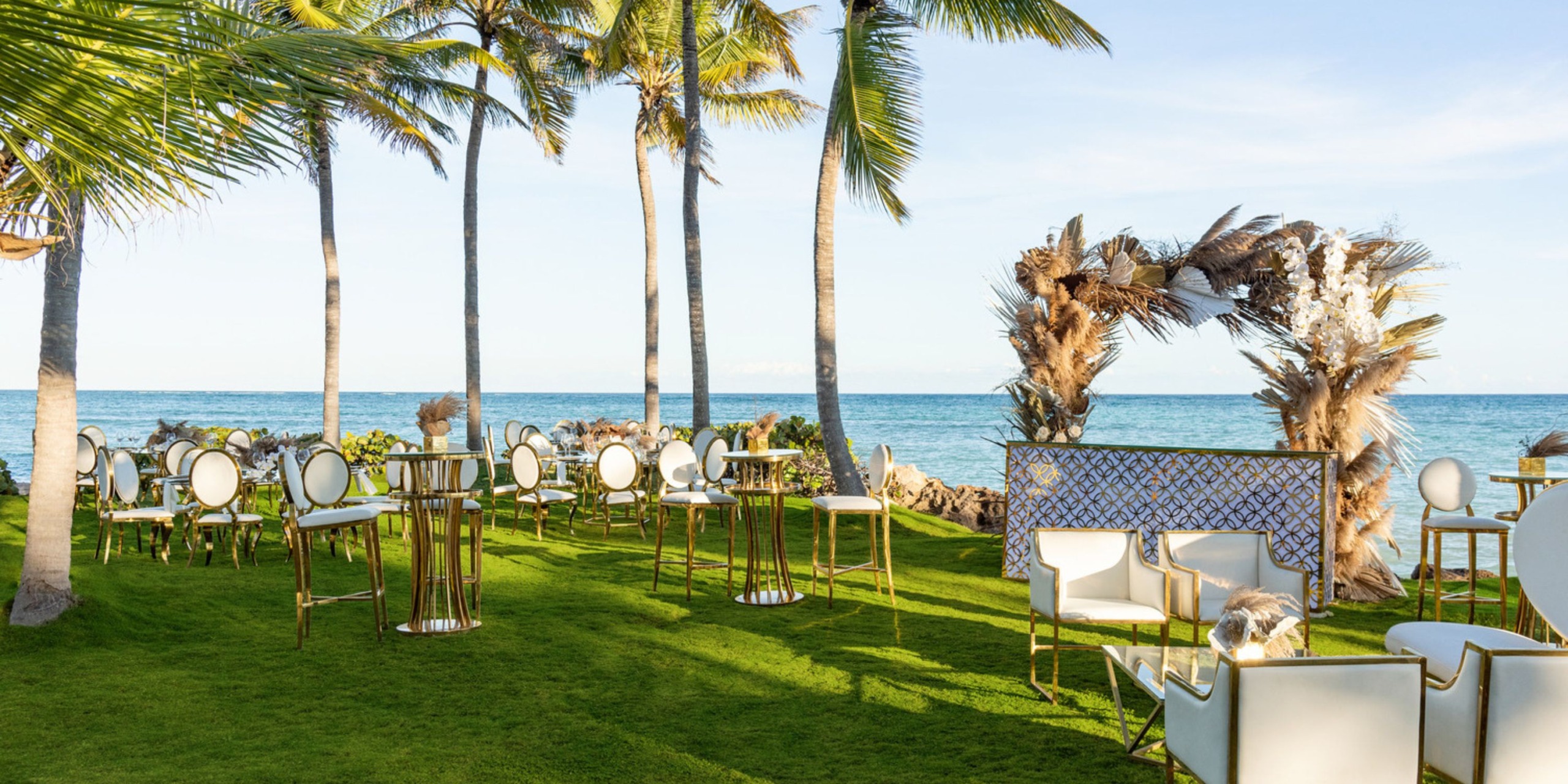The Sanctuary Cap Cana offers an outdoor wedding setup by the ocean, complete with palm trees, white and gold chairs, elegant tables, and a beautifully decorated arch on a grassy area.