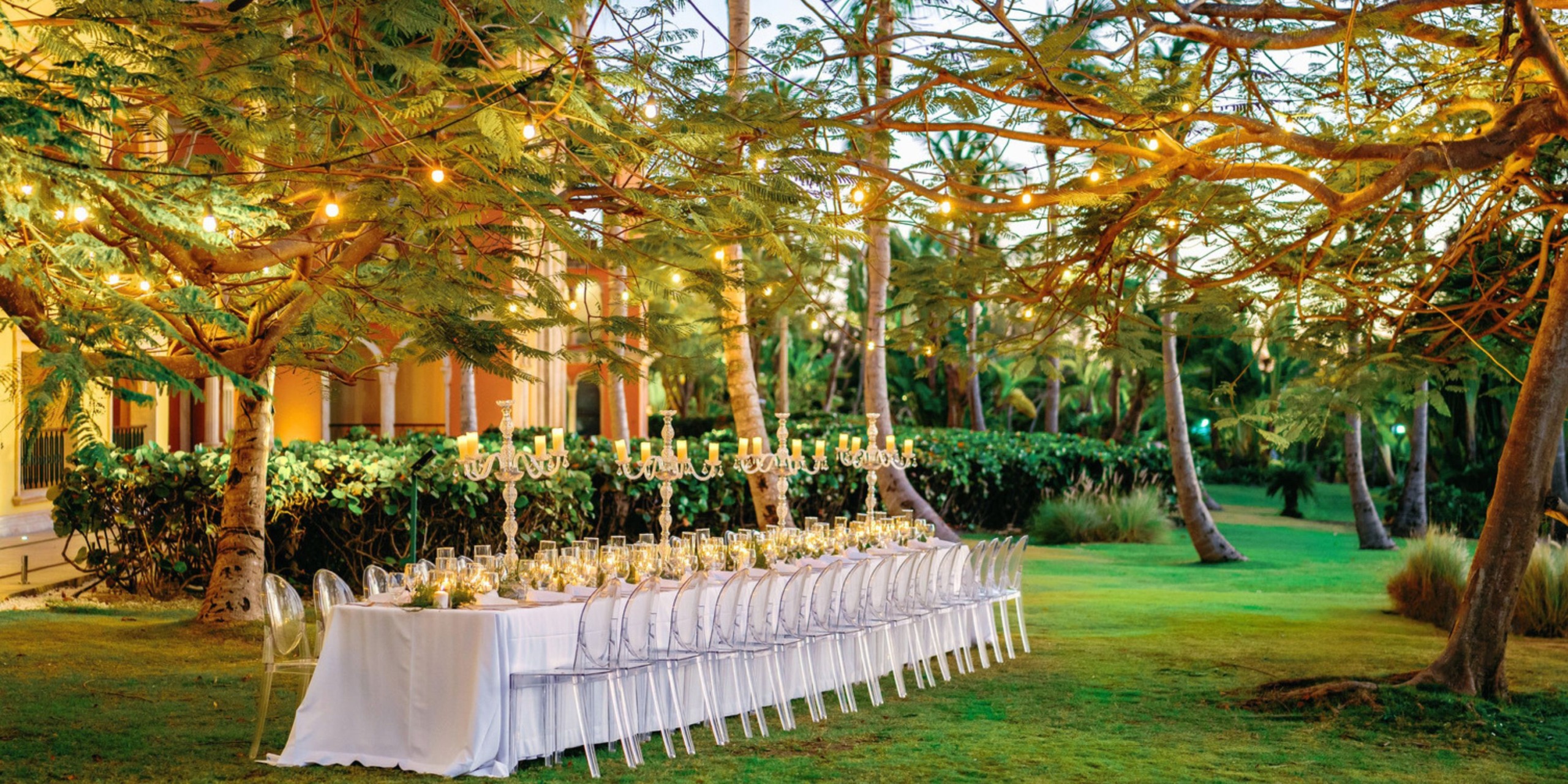 The Sanctuary Cap Cana presents an outdoor dinner arrangement featuring a long white table, transparent chairs, and sophisticated table settings beneath string lights in a garden ambiance.