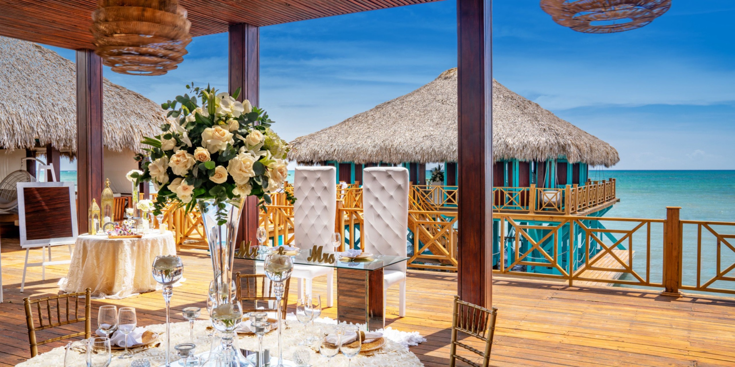 An enchanting wedding setup at Sanctuary Cap Cana, located on a wooden deck by the ocean, boasting sophisticated table arrangements adorned with white flowers and a picturesque thatched-roof building in the background.