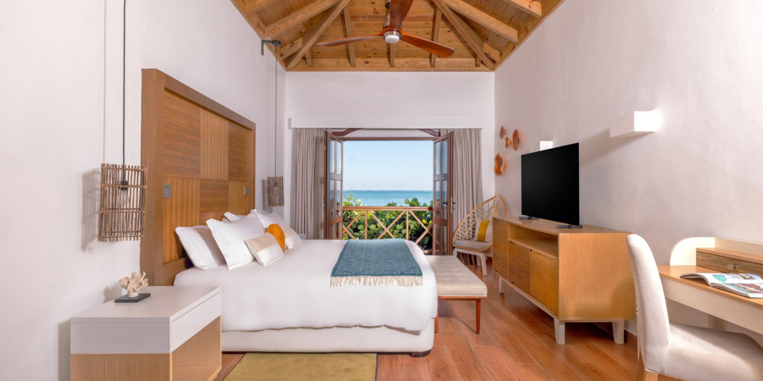 The Sanctuary Cap Cana features a contemporary hotel room equipped with a spacious bed, television, and elegant wooden furnishings. Through an open sliding door, you'll find a balcony that offers stunning views of the ocean and lush greenery.