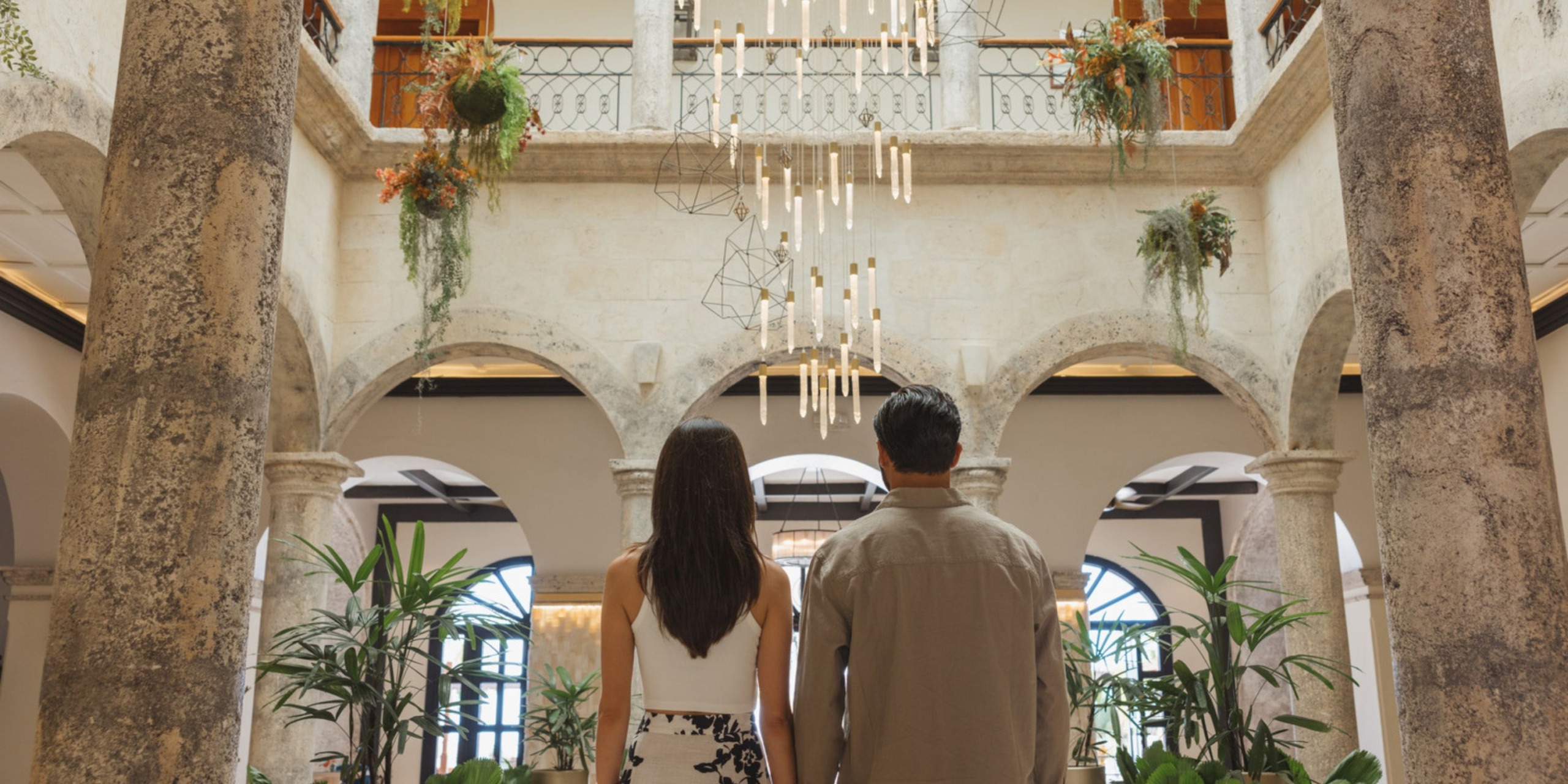 A couple stands in the grand atrium of Sanctuary Cap Cana, filled with lush greenery and arched doorways, gazing up at a modern chandelier.