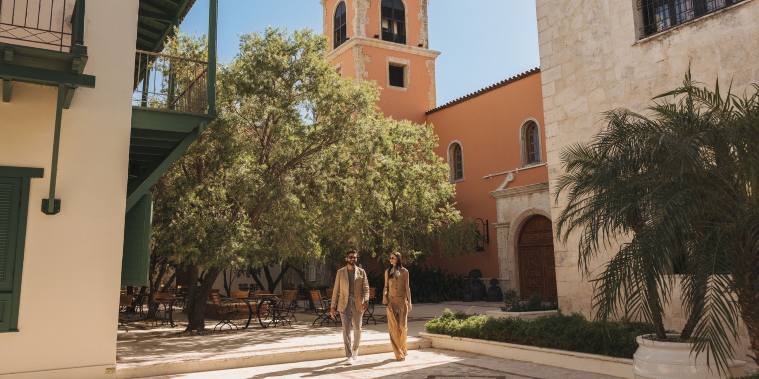 Two people stroll through the sunlit courtyard of Sanctuary Cap Cana, surrounded by outdoor seating, lush trees, and historic architecture.