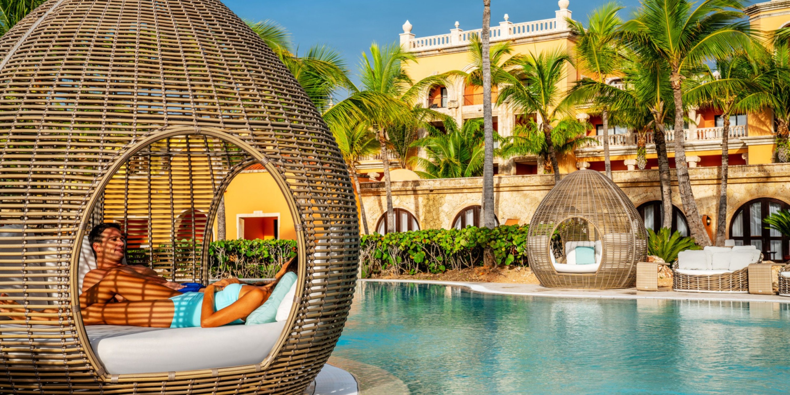 A person relaxes in a wicker cabana by a pool at Sanctuary Cap Cana, surrounded by palm trees and with a yellow building in the background.