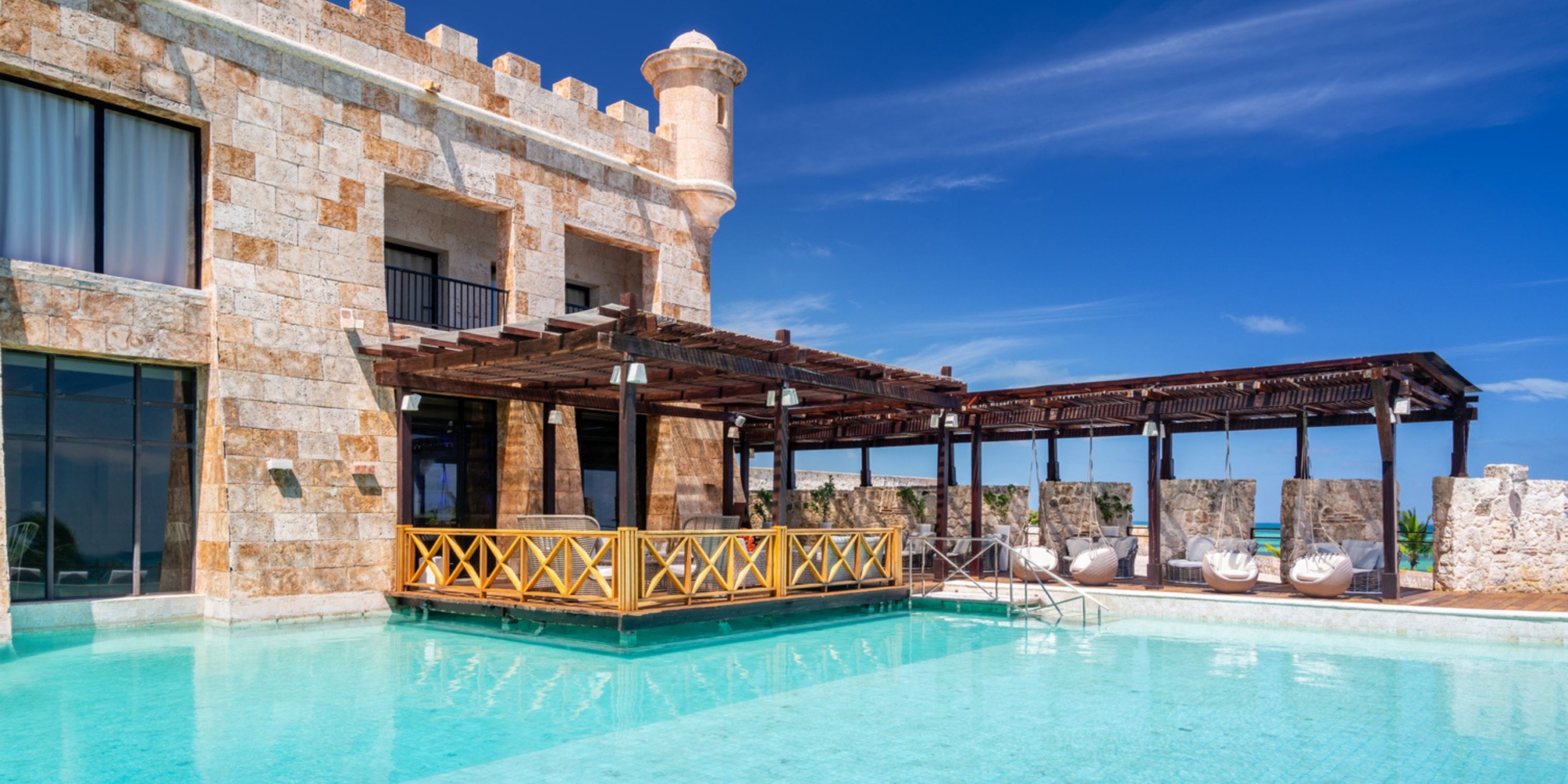 Discover the Sanctuary Cap Cana, a luxurious stone villa featuring a balcony that overlooks a crystal-clear blue swimming pool. Adding to its charm, a wooden pergola offers shade over part of the pool. Enjoy the beauty of clear and sunny skies in this serene setting.
