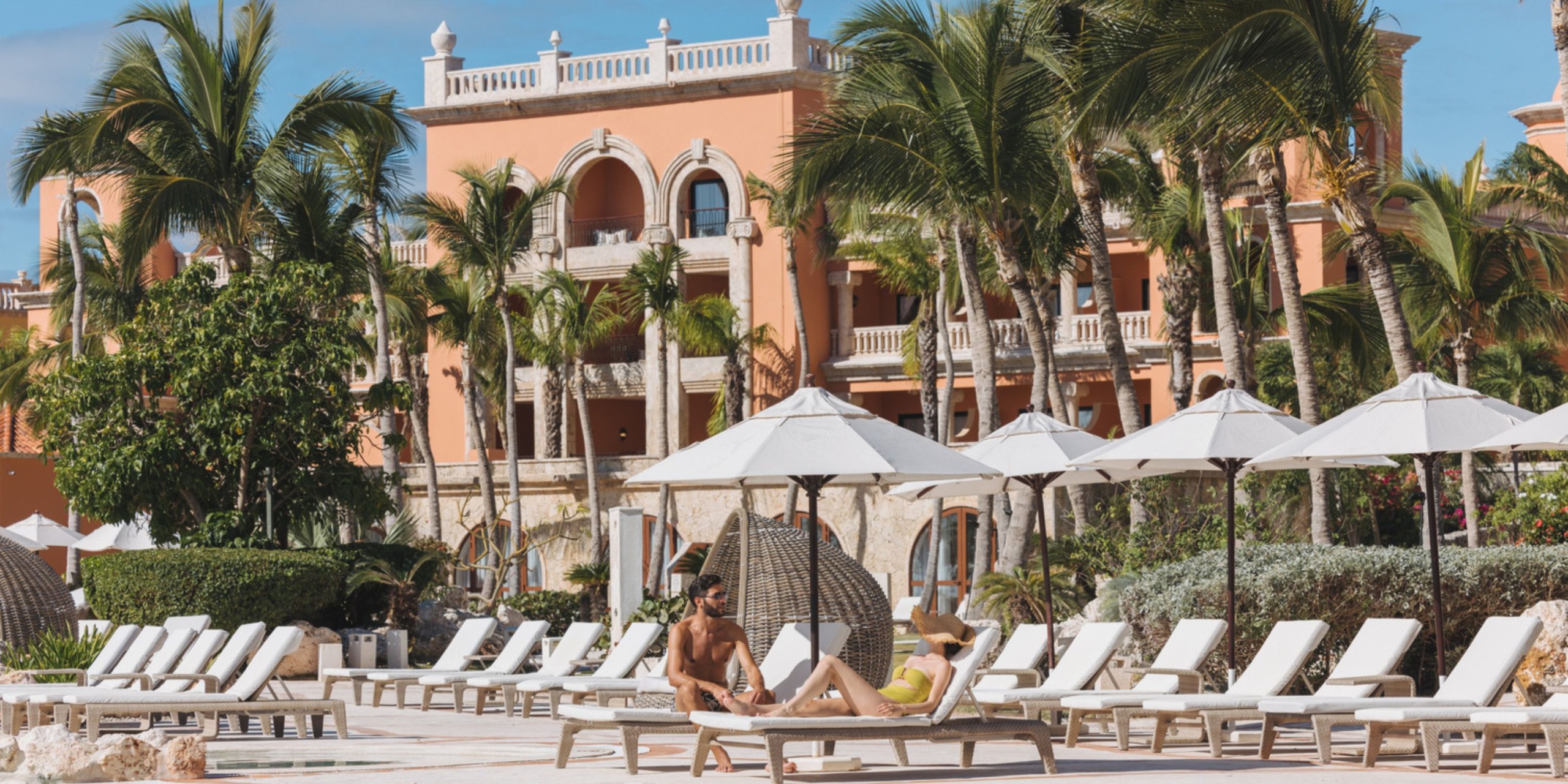 Guests unwind on lounge chairs by the pool shaded by white umbrellas at Sanctuary Cap Cana, with a large building and palm trees framing the backdrop.
