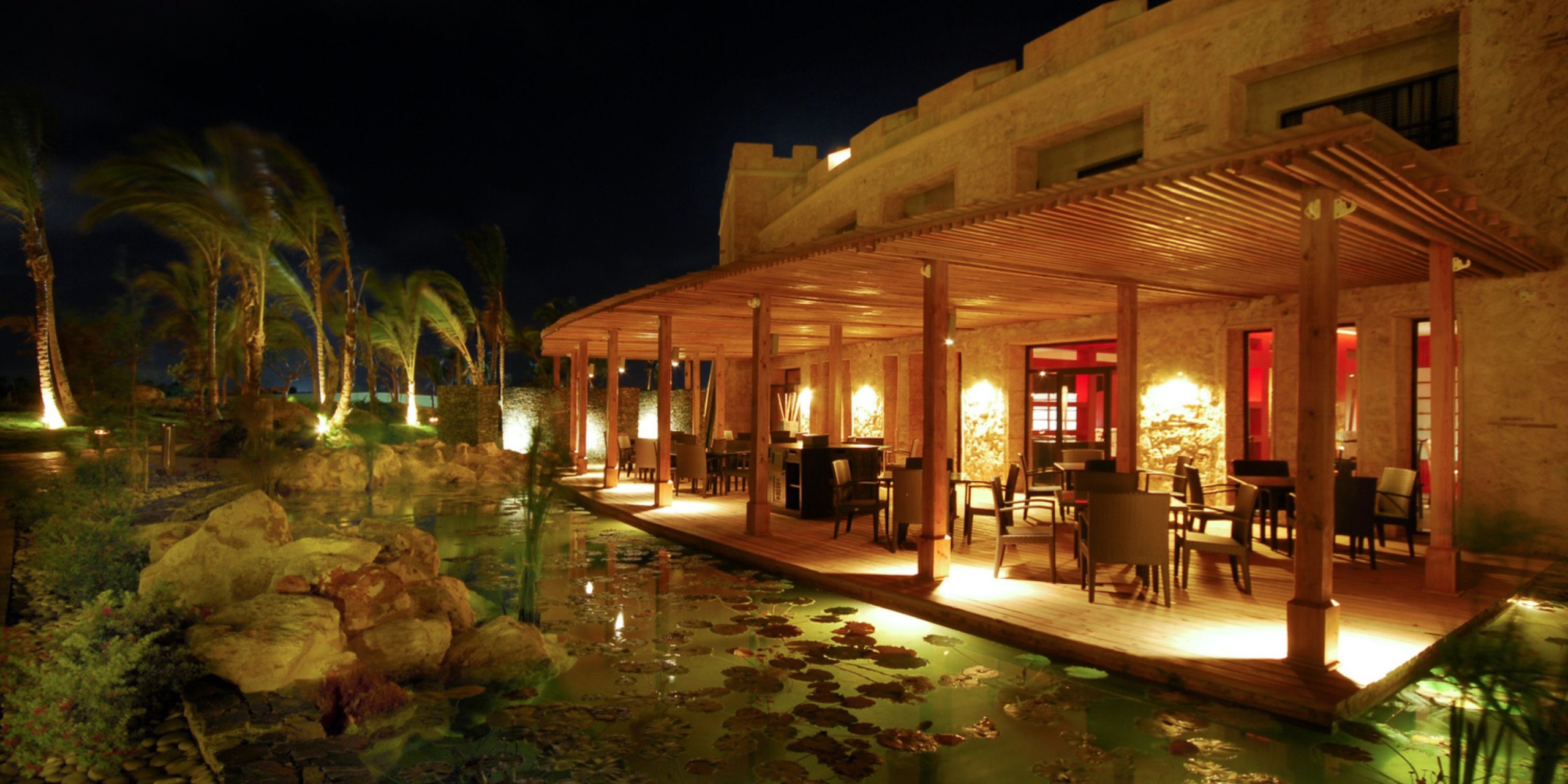 The outdoor patio at Sanctuary Cap Cana is illuminated under the night sky, featuring wooden pergolas, tables, and chairs, with nearby palm trees and a pond adorned with lily pads.