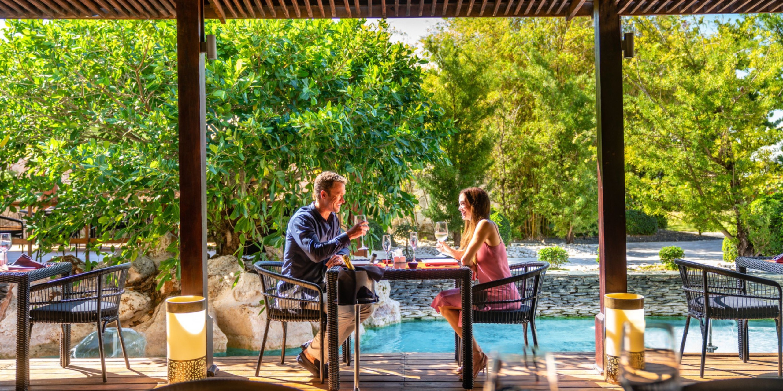 A couple sits at a table, enjoying a meal and wine outdoors by the pool at Sanctuary Cap Cana, enveloped by trees and natural light.