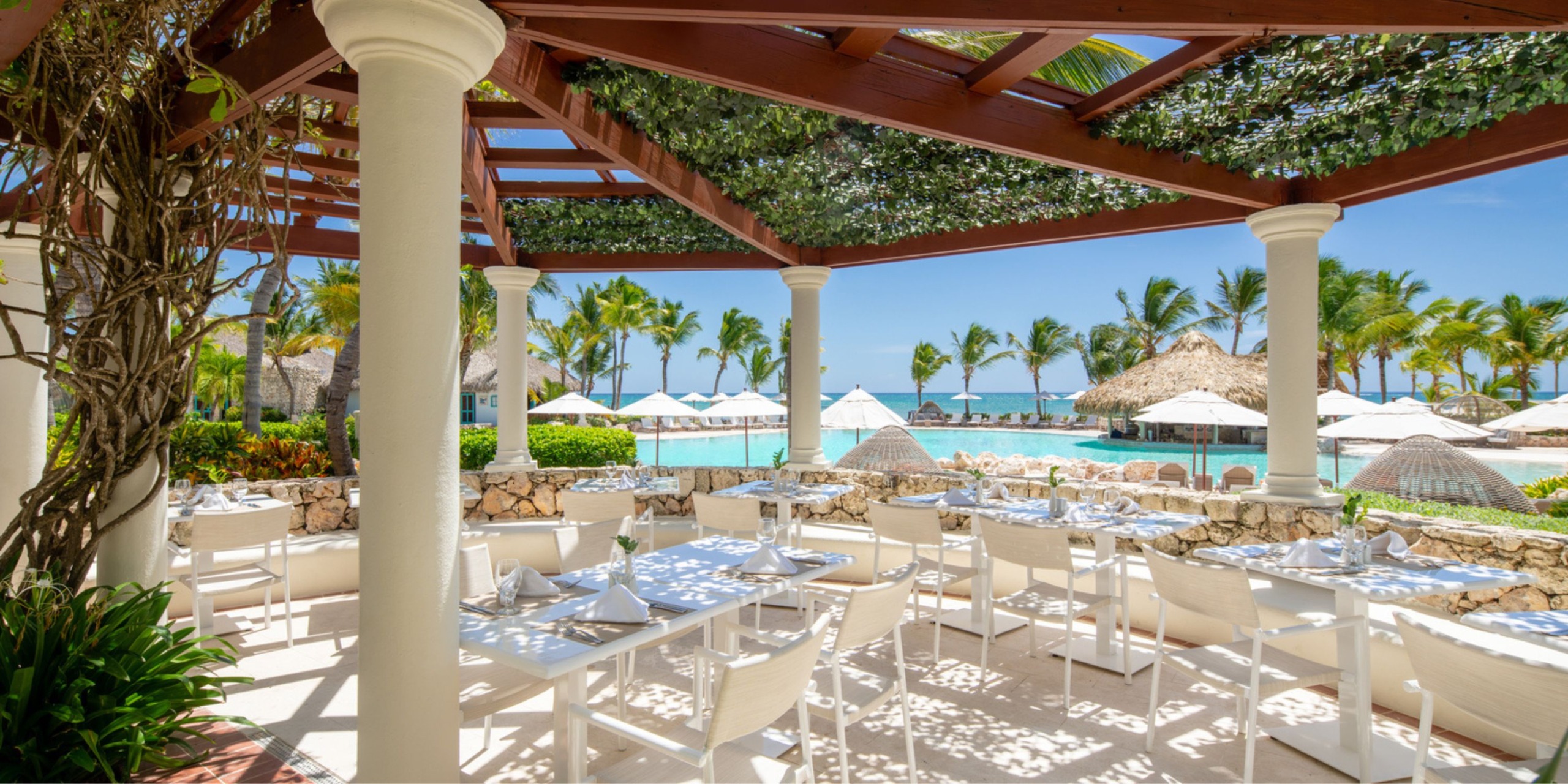 The Sanctuary Cap Cana features an outdoor dining area with white tables and chairs under a wooden pergola, offering a stunning view of the pool and beach, complete with palm trees and umbrellas in the background.