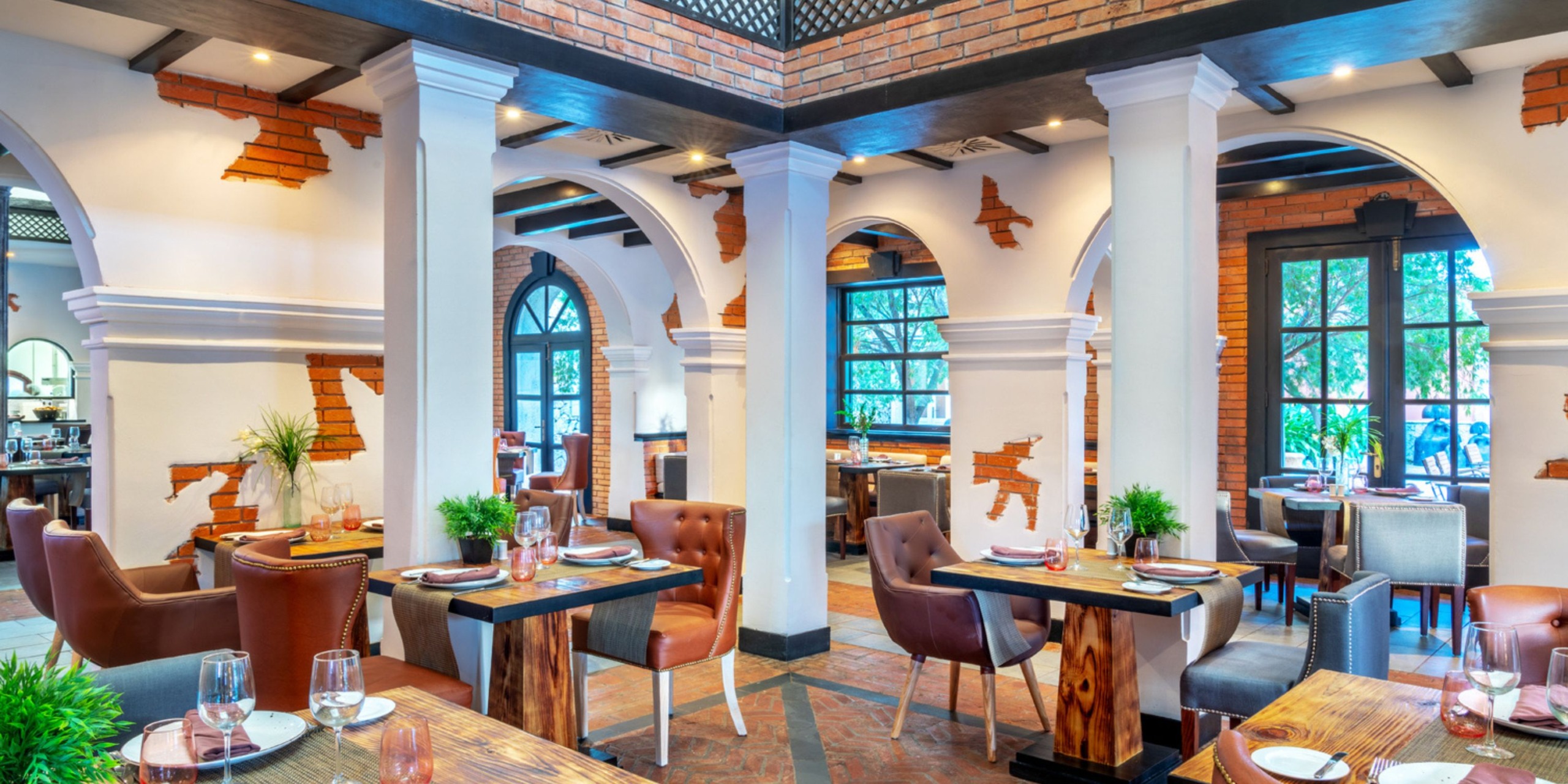 The Sanctuary Cap Cana offers a spacious restaurant interior featuring brick and white arched walls, wooden tables, and a mix of leather and fabric chairs, all illuminated by natural light streaming through large windows.