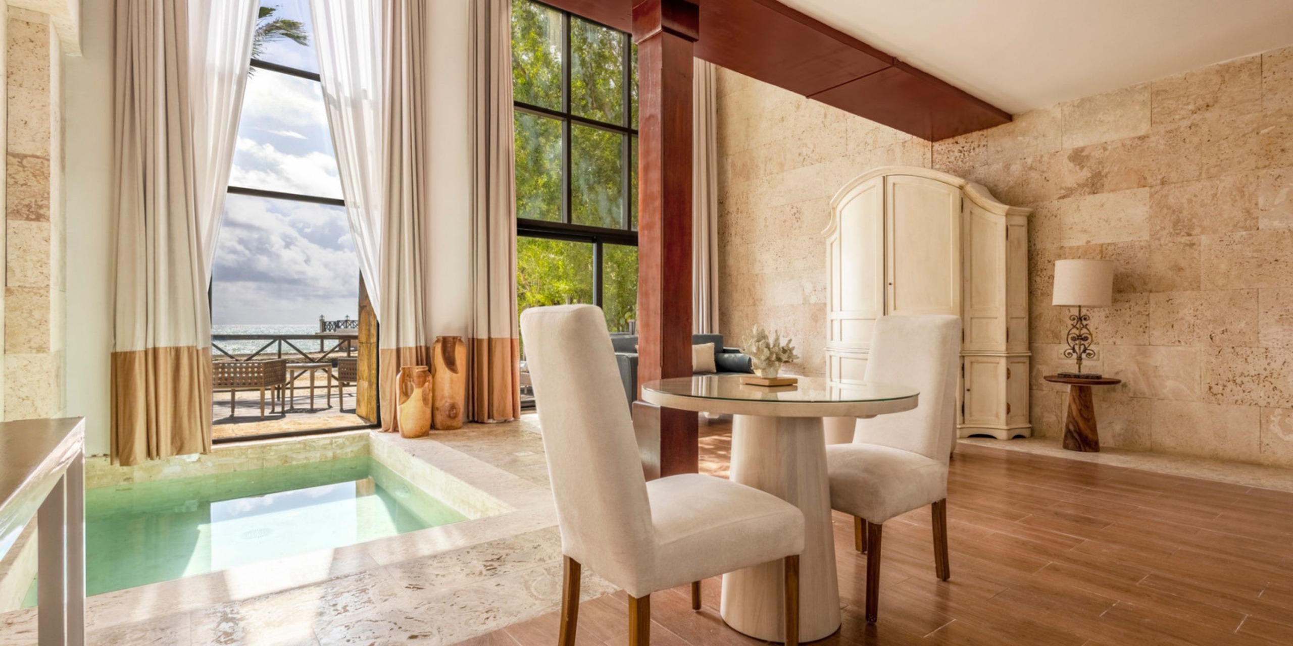 The Sanctuary Cap Cana features a bright room complete with a small indoor pool, a round table with two chairs, large windows, and an armoire. Through the open curtains, you can enjoy the view of a balcony overlooking the sea.