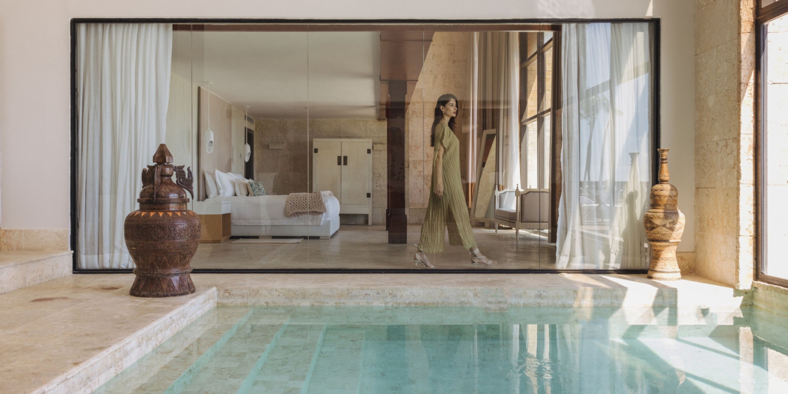 A person in a long dress walks into the modern, glass-walled space of Sanctuary Cap Cana, which overlooks a small pool and features stone accents and large decorative vases nearby.