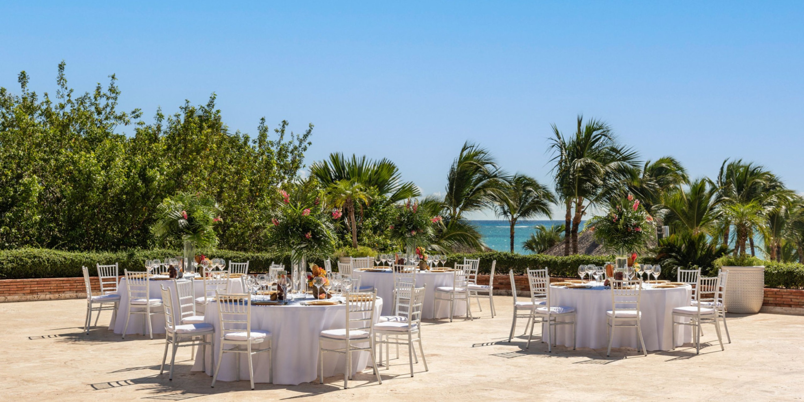 Sanctuary Cap Cana's outdoor dining area features round tables with white chairs and floral centerpieces, set against a backdrop of palm trees and the ocean beneath a clear blue sky.