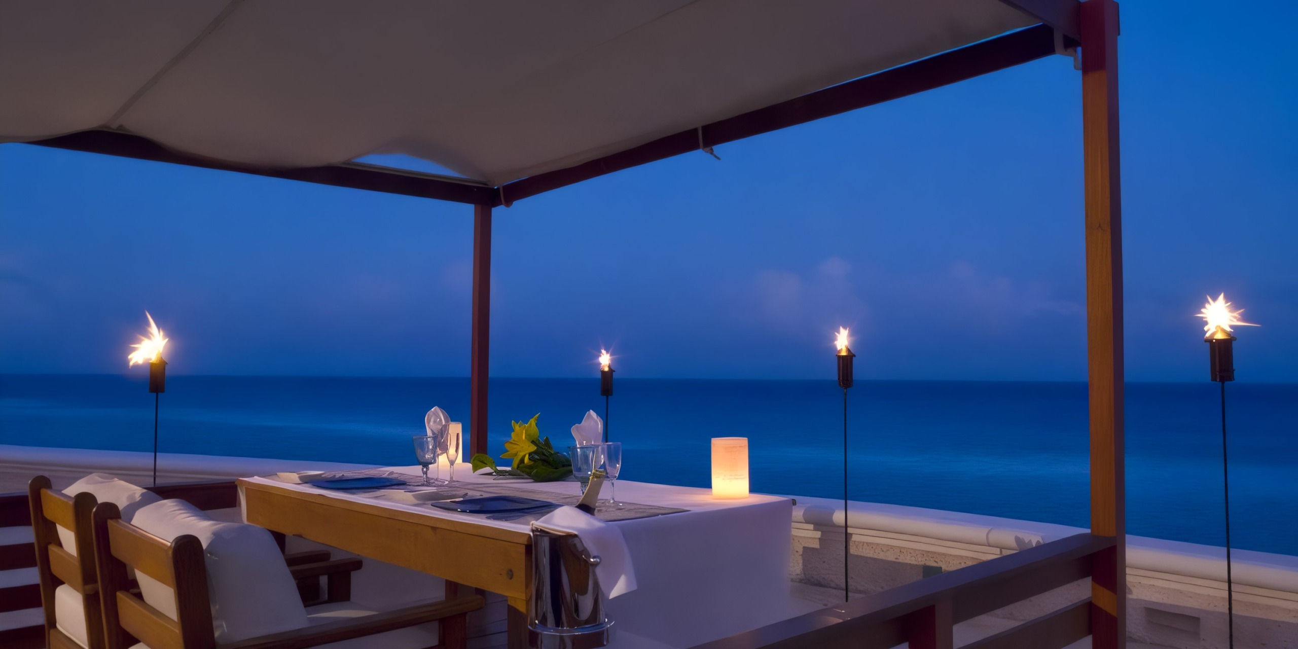 A Sandos Cancun Lifestyle Resort dining experience for two under a canopy, complete with lit torches and a candle, overlooking the calm blue ocean at dusk.