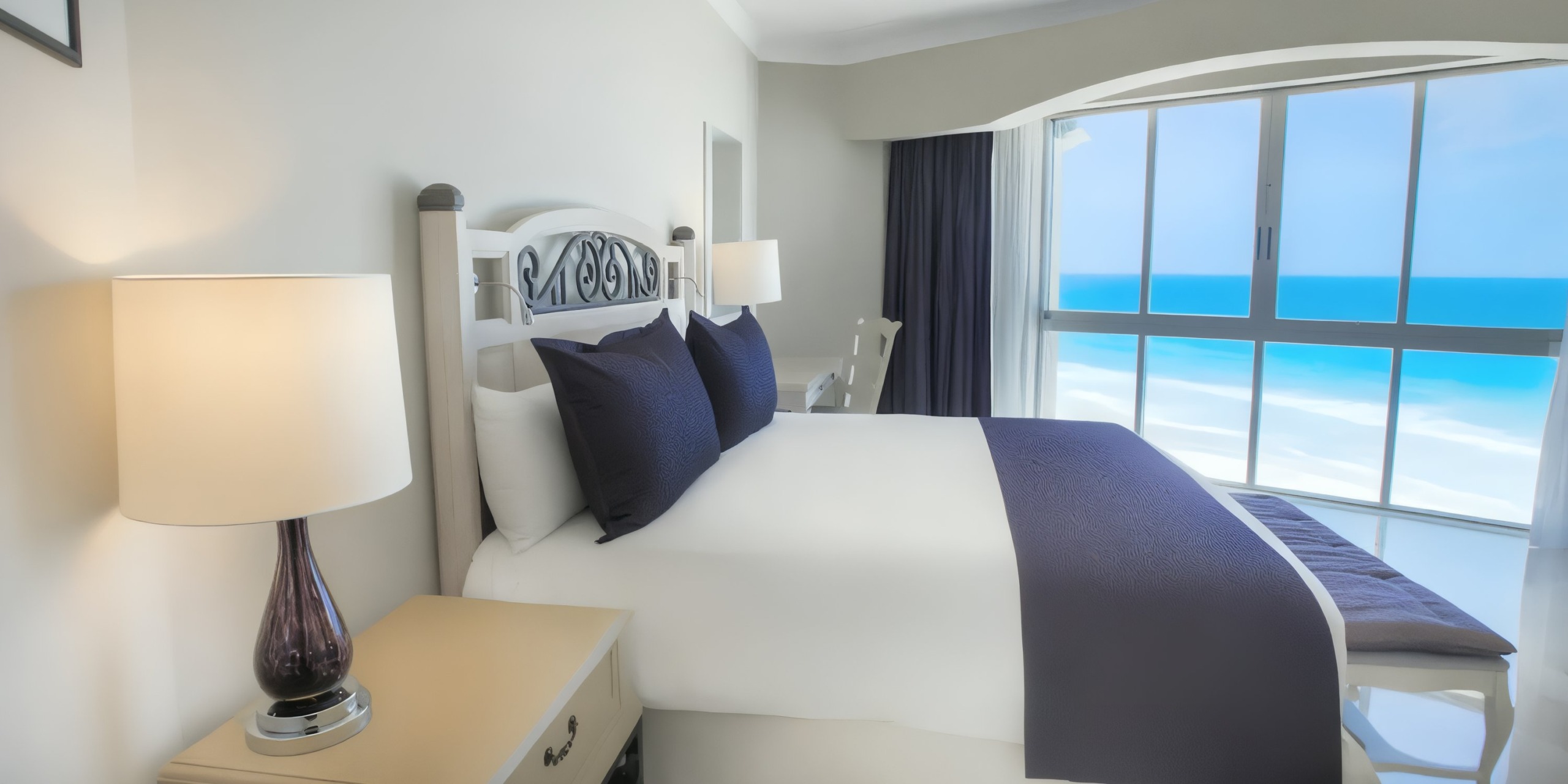 A bedroom at the Sandos Cancun Lifestyle Resort features a large bed, a bedside lamp, and an ocean view through the window. The white and navy bedding contrasts beautifully with the light-colored walls and floors.