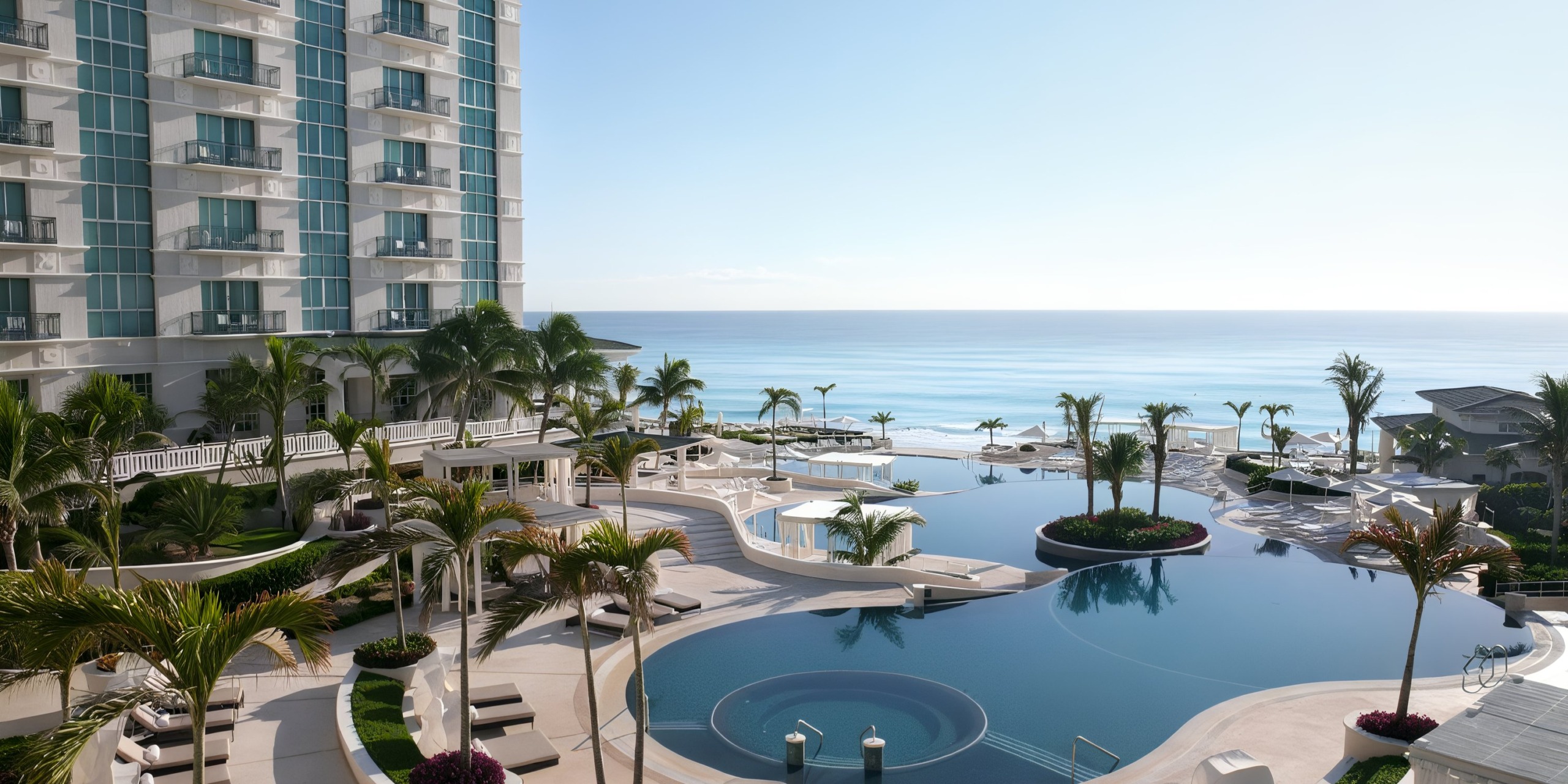 The Sandos Cancun Lifestyle Resort boasts a stunning curved infinity pool, encircled by palm trees and sun loungers, offering an unobstructed view of the ocean. A multi-story hotel building is situated to the left.