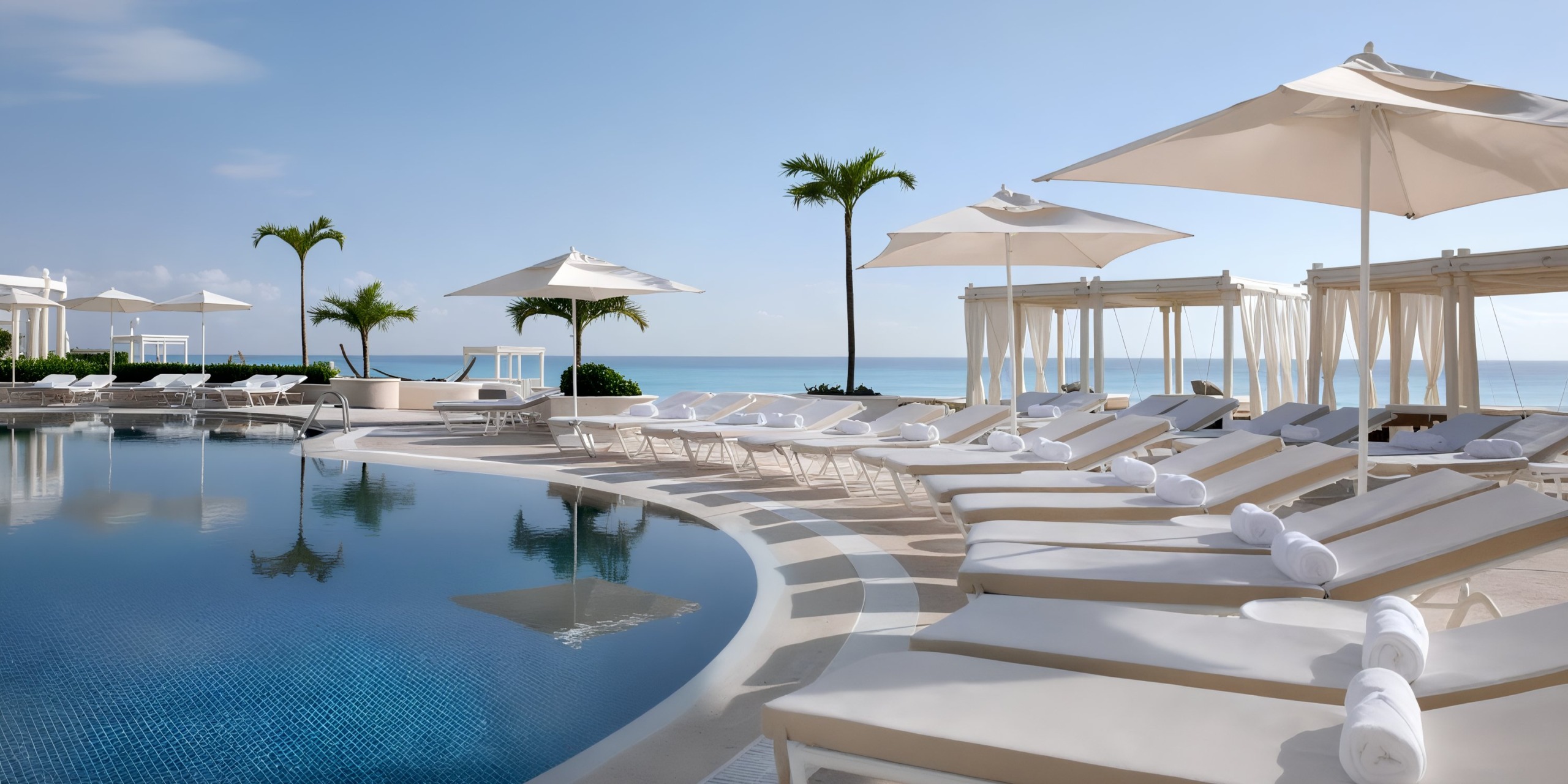 A tranquil poolside setting at Sandos Cancun Lifestyle Resort features lounge chairs, umbrellas, and cabanas with a stunning view of the ocean under a clear blue sky. Two palm trees stand elegantly near the water.