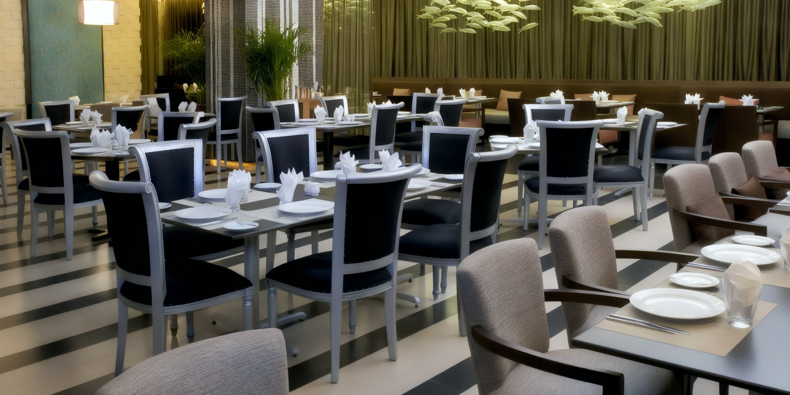 The Sandos Cancun Lifestyle Resort features a modern dining setting with neatly arranged tables and chairs, elegantly set with white tablecloths, plates, and folded napkins. Green plants and decorative lighting create an inviting atmosphere in the background.