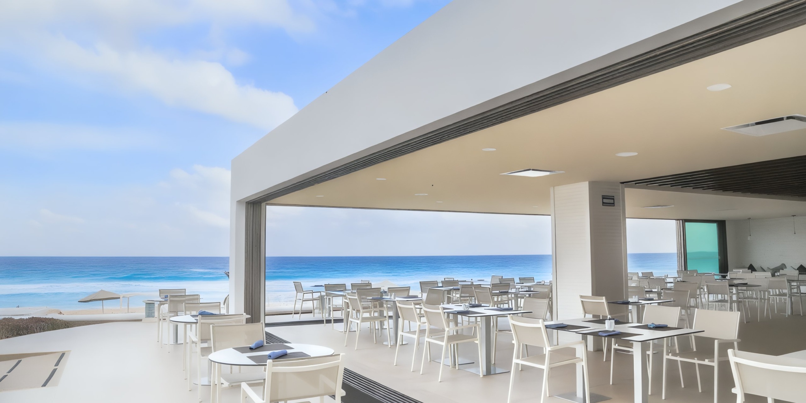 Experience the outdoor seaside dining area at Sandos Cancun Lifestyle Resort, featuring white tables and chairs under a sleek open-air structure, all with stunning views of the clear blue ocean and sky.