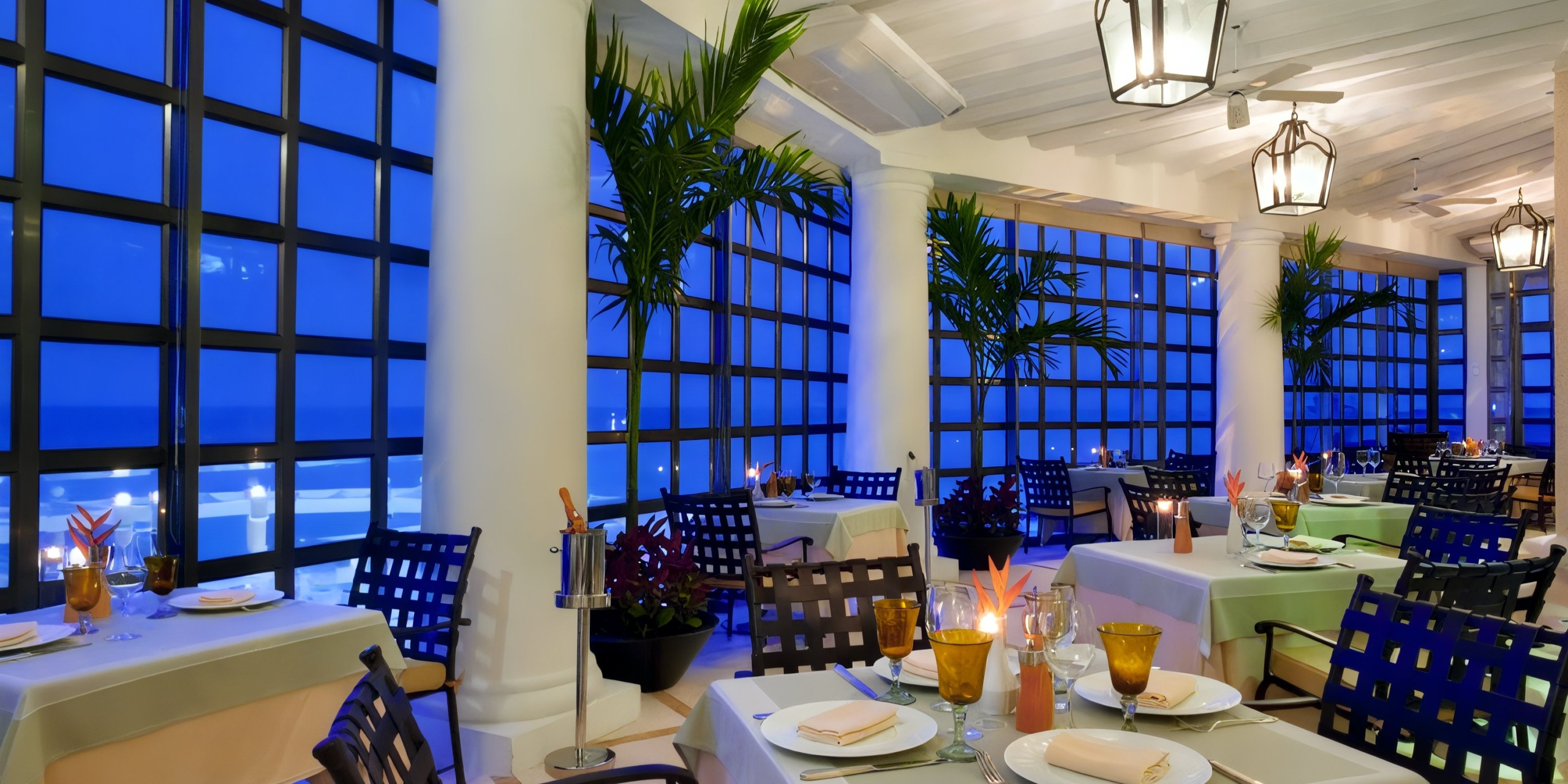 The Sandos Cancun Lifestyle Resort features a picturesque seaside restaurant with neatly arranged tables, adorned with lush plants and elegant lanterns, offering breathtaking views of the ocean through expansive windows at dusk.