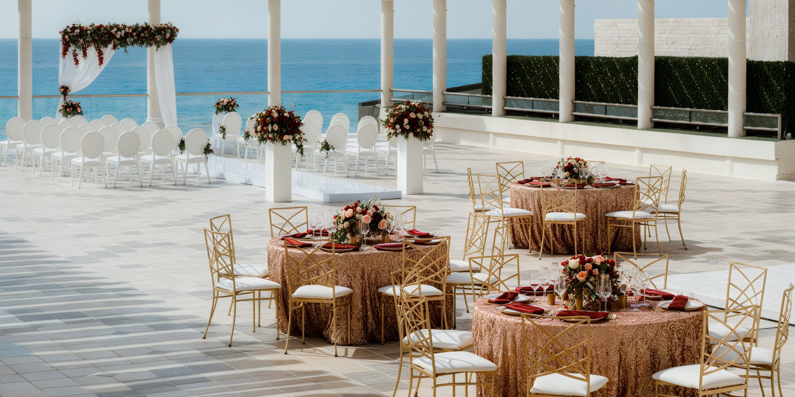 At Sandos Cancun Lifestyle Resort, envision an outdoor wedding setup featuring round tables adorned with pink tablecloths, elegant floral centerpieces, and sophisticated gold chairs. The ceremonial area includes a charming arch overlooking the ocean under a clear sky.