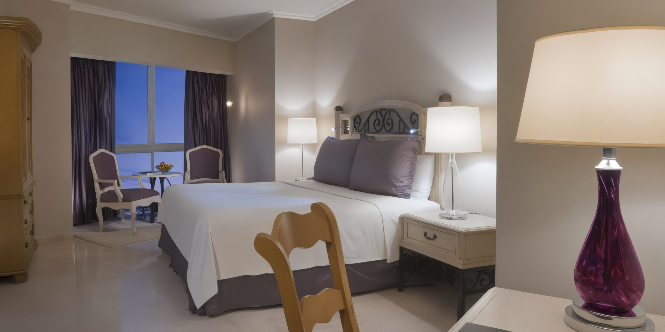 Sandos Cancun Lifestyle Resort offers a hotel room with a neatly made double bed, two bedside lamps, a small table with two chairs by the window, and a wooden chair in the foreground.