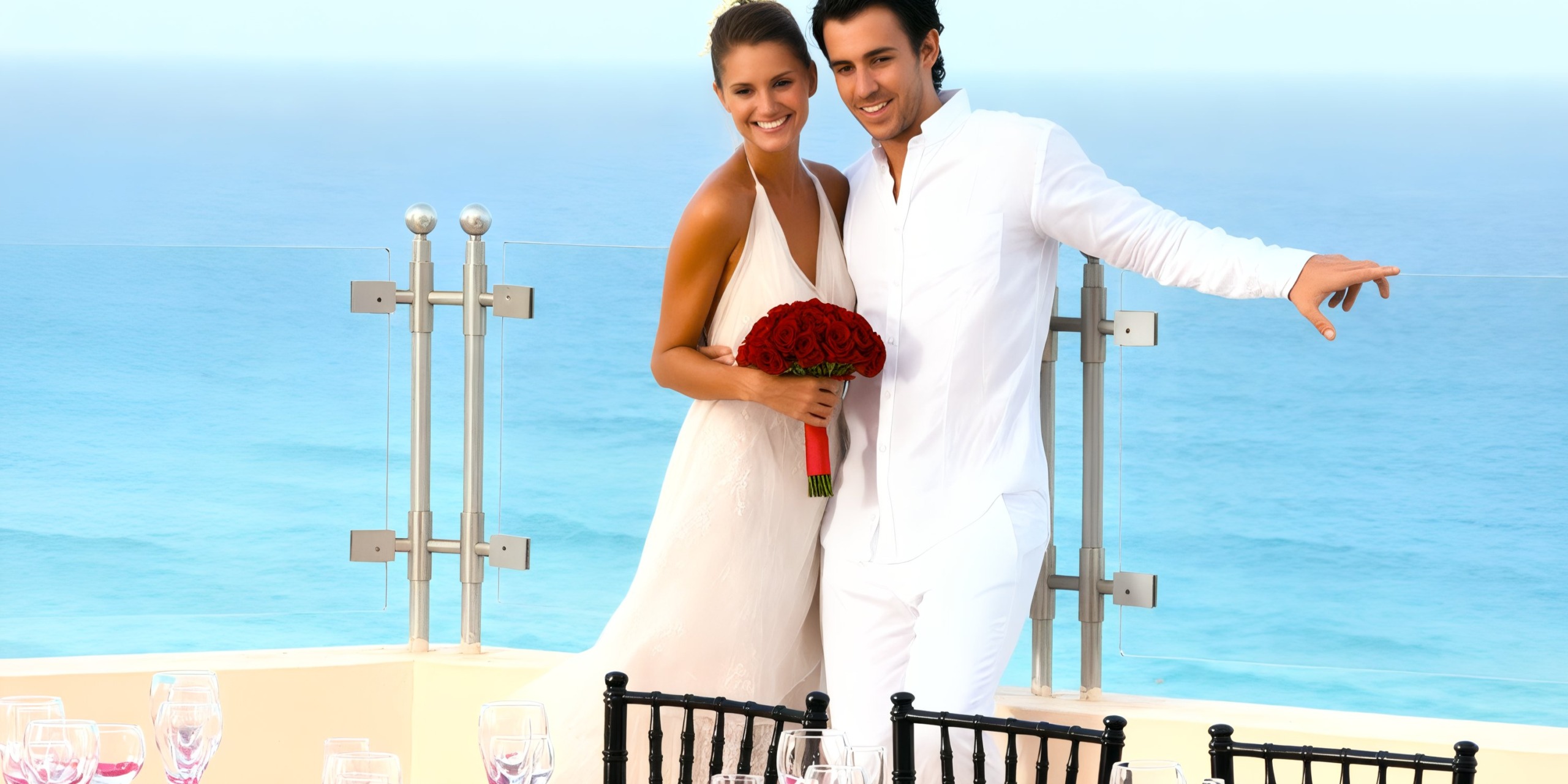 A couple dressed in white poses for a photo on a terrace at Sandos Cancun Lifestyle Resort, overlooking the ocean. The woman holds a bouquet of red roses, and the setting suggests a celebration or event.