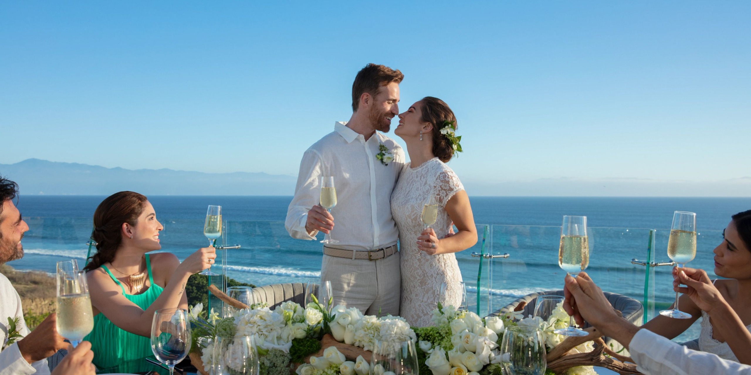 Dressed in wedding attire, a couple embraces with champagne glasses in hand, enjoying the seaside view at Secrets Bahia Mita. Meanwhile, guests at a nearby table raise their glasses, with the ocean serving as a stunning backdrop.