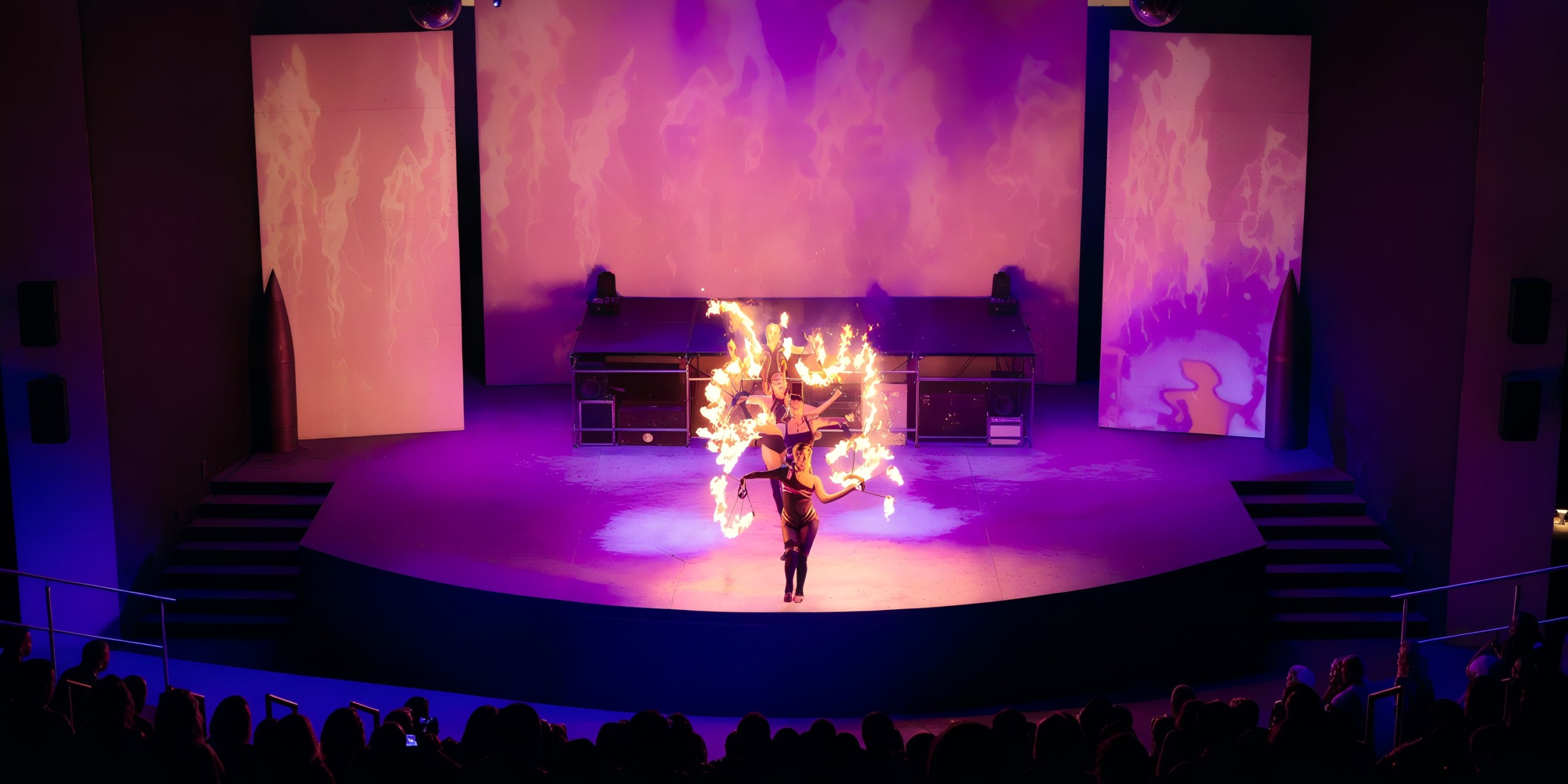 Under mesmerizing purple lighting, a performer expertly maneuvers flaming hoops on stage against a backdrop of projected flames, captivating the audience at Secrets Bahia Mita.