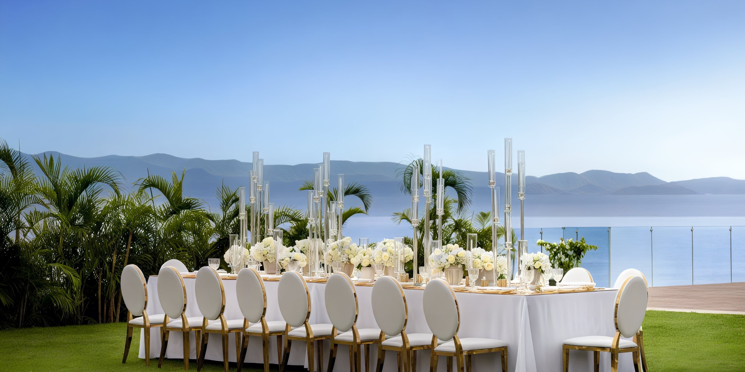 Create a picturesque outdoor dining setup featuring a long table with white chairs, adorned with floral centerpieces and candlesticks, offering a stunning view of the body of water and distant mountains at Secrets Bahia Mita.