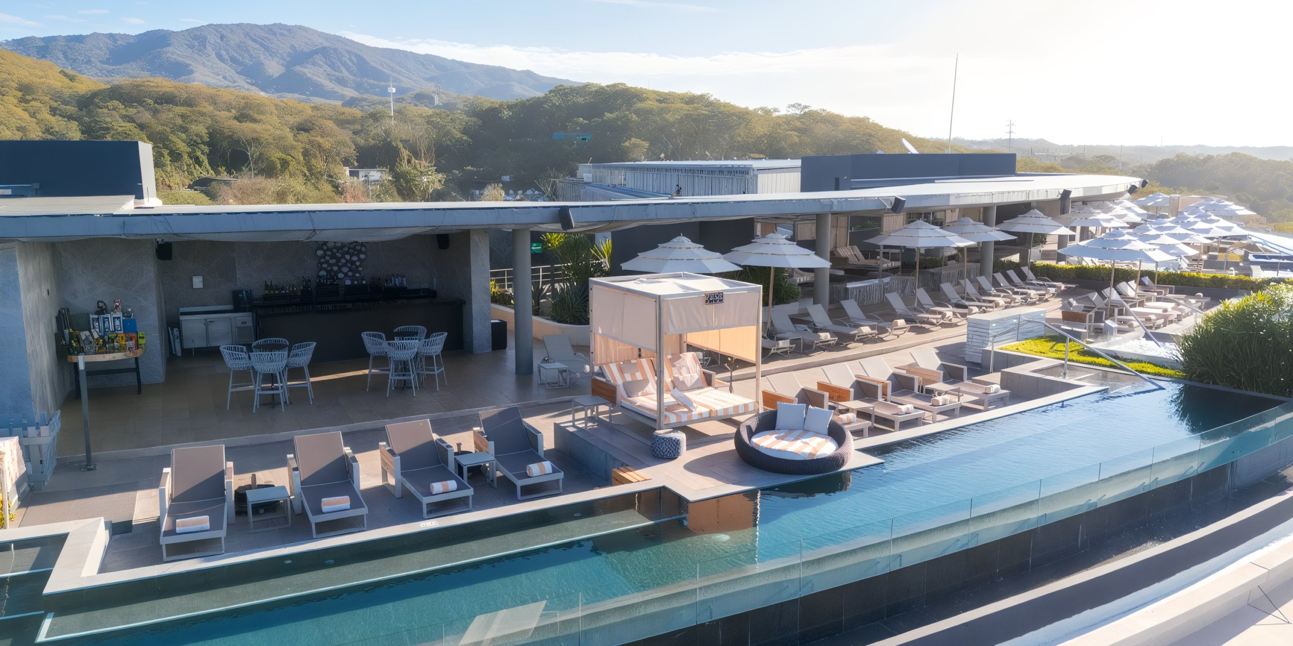 The rooftop pool area at Secrets Bahia Mita features lounge chairs, umbrellas, and a gazebo, all set against the backdrop of hills and trees under a clear sky.