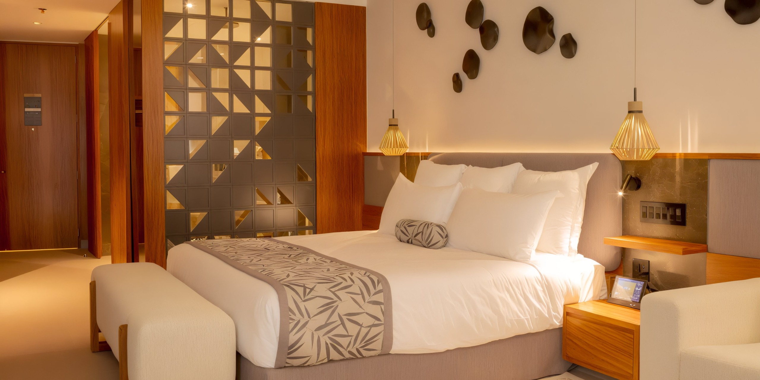 The Secrets Bahia Mita hotel room features a large bed adorned with decorative pillows and a patterned throw, complemented by warm lighting, geometric wall art, and a sleek wooden headboard with integrated nightstands.
