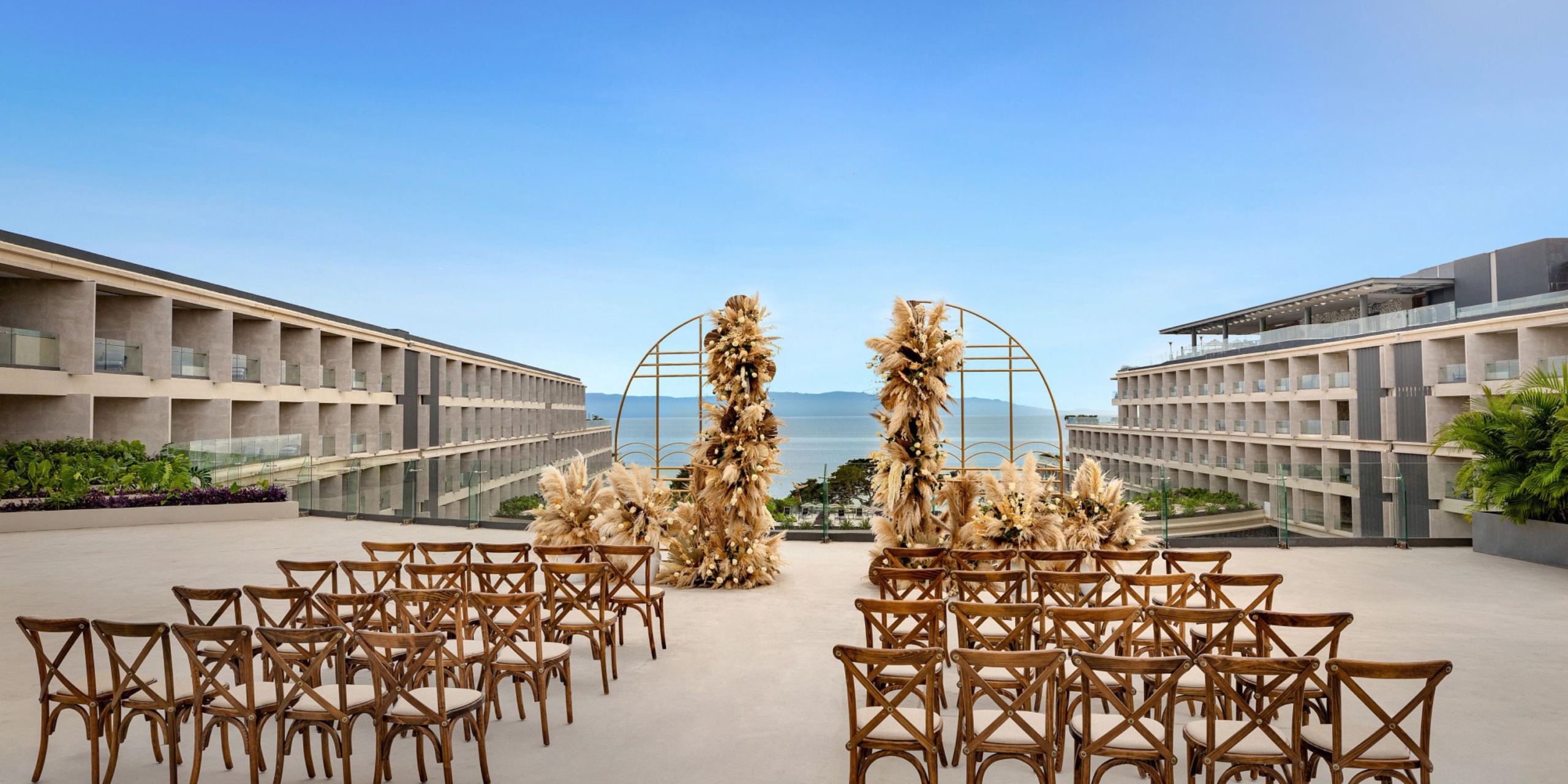 Set up for an outdoor wedding at Secrets Bahia Mita features wooden chairs arranged to face an arch adorned with dried flowers, overlooking a reflective surface and ocean view, and flanked by modern buildings.
