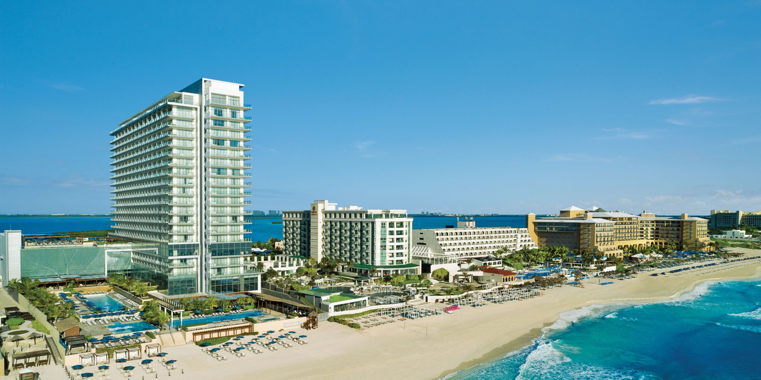 Aerial view of Secrets the Vine Cancun, a beachfront resort featuring high-rise structures along a sandy coastline with clear blue waters under a sunny sky.